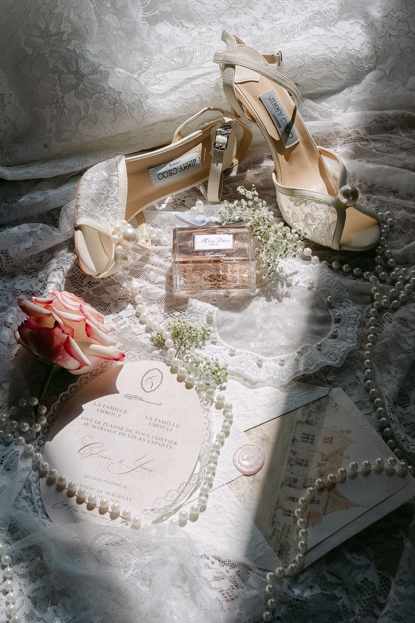 Elegant bridal flat lay with Jimmy Choo heels, Dior perfume, pearl necklace, roses, and wedding invitations on lace fabric.