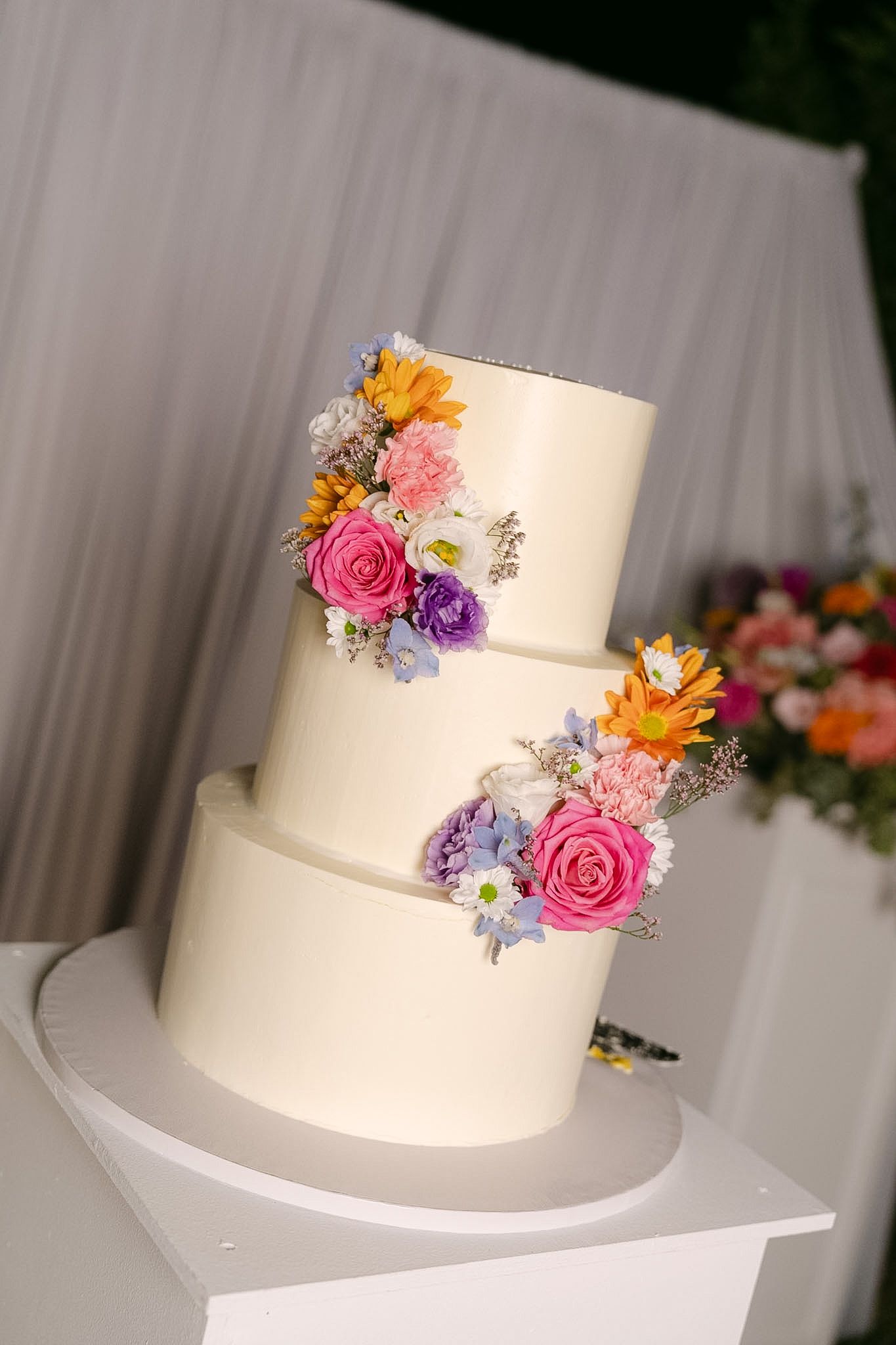 Elegant two-tier white wedding cake adorned with colorful fresh flowers including pink roses and purple blooms.