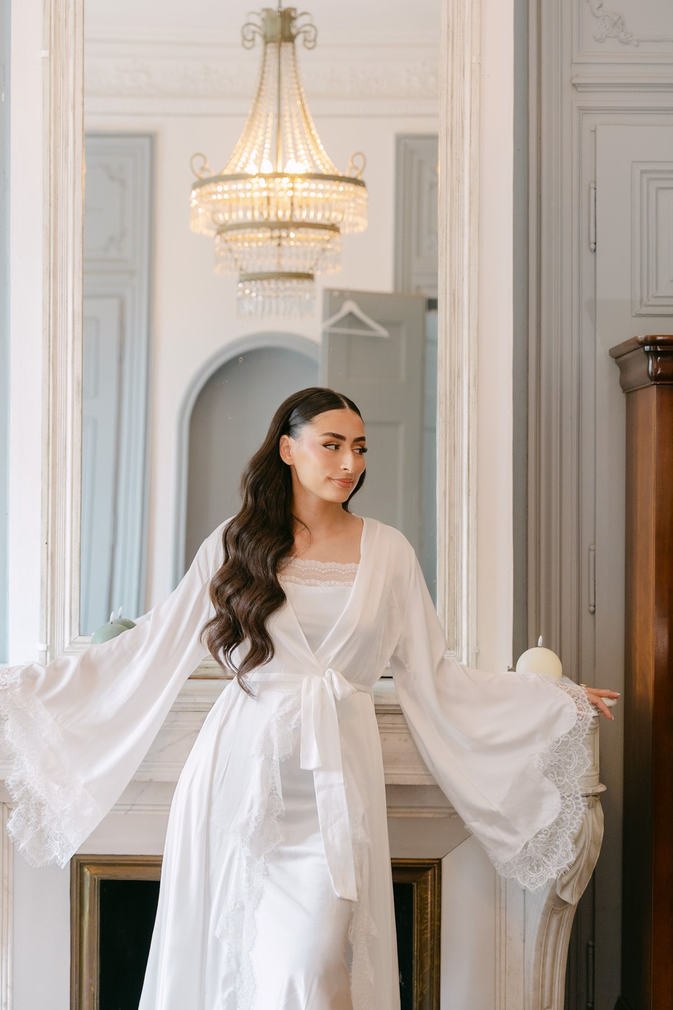 Bride in white lace robe with flowing sleeves stands by fireplace under crystal chandelier in elegant room.