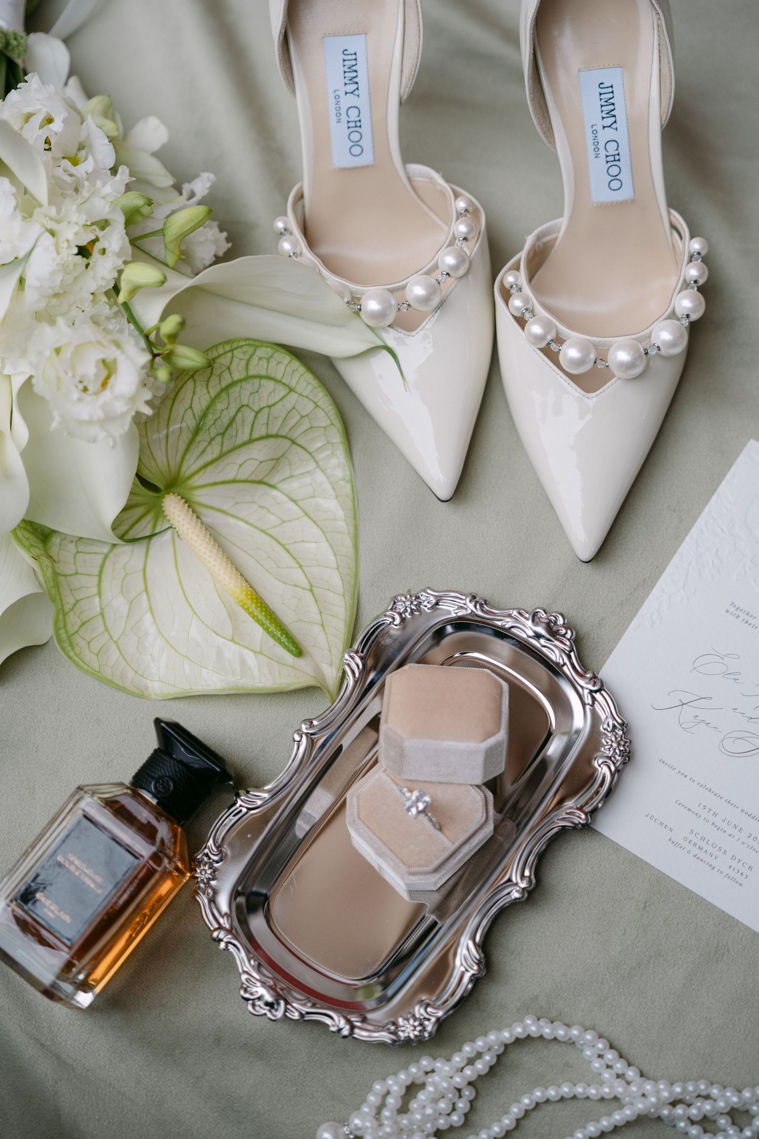 Elegant bridal flat lay featuring Jimmy Choo pearl heels, ring box on silver tray, perfume bottle, pearl necklace, and white floral bouquet.