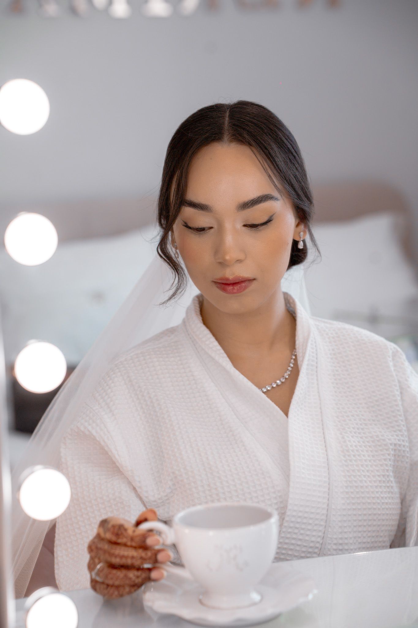 Beautiful bride in white robe and veil holding teacup at vanity mirror during wedding morning preparation.