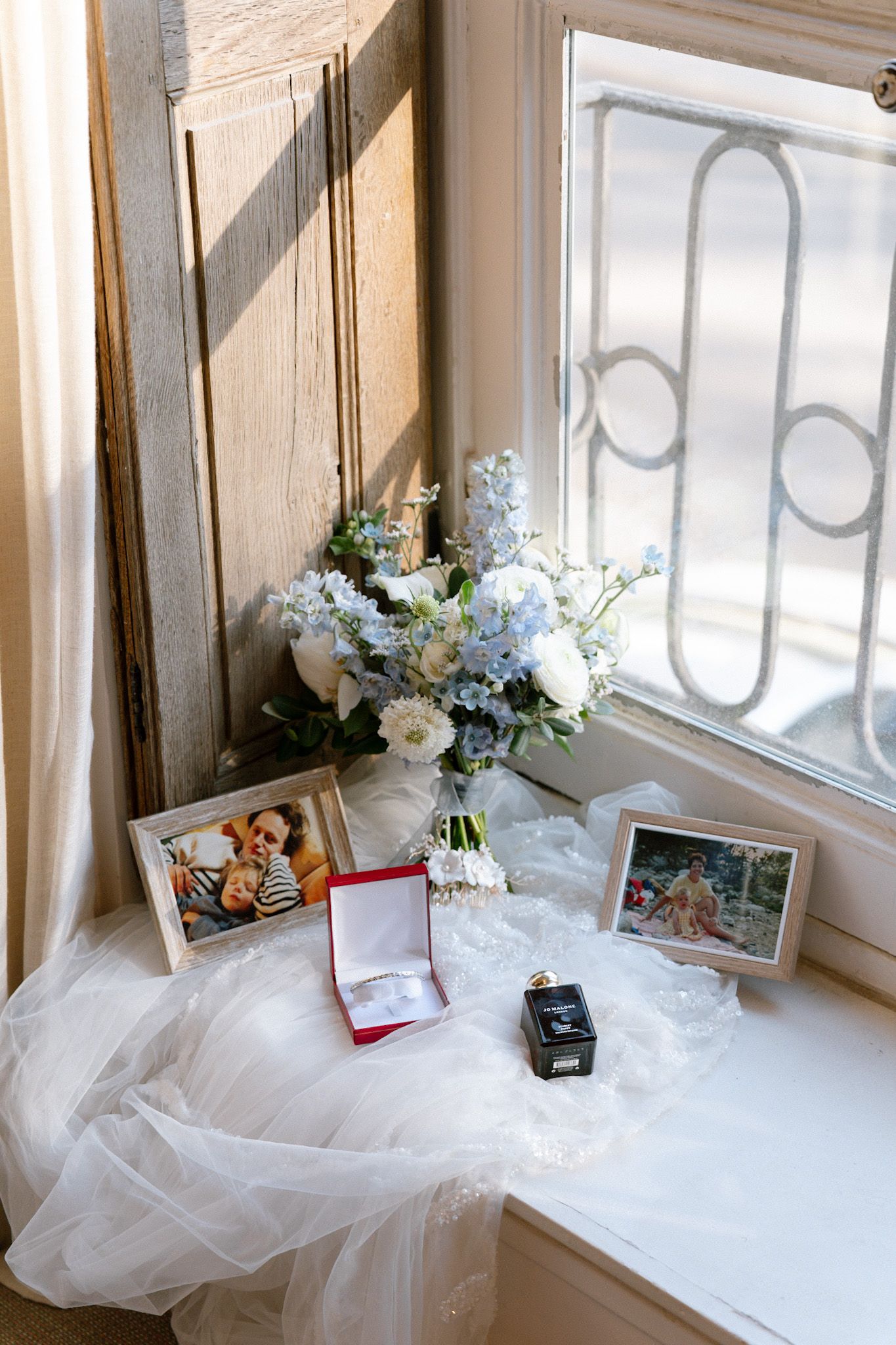 Wedding details on windowsill with floral bouquet, framed photos, and ring boxes on white veil fabric.