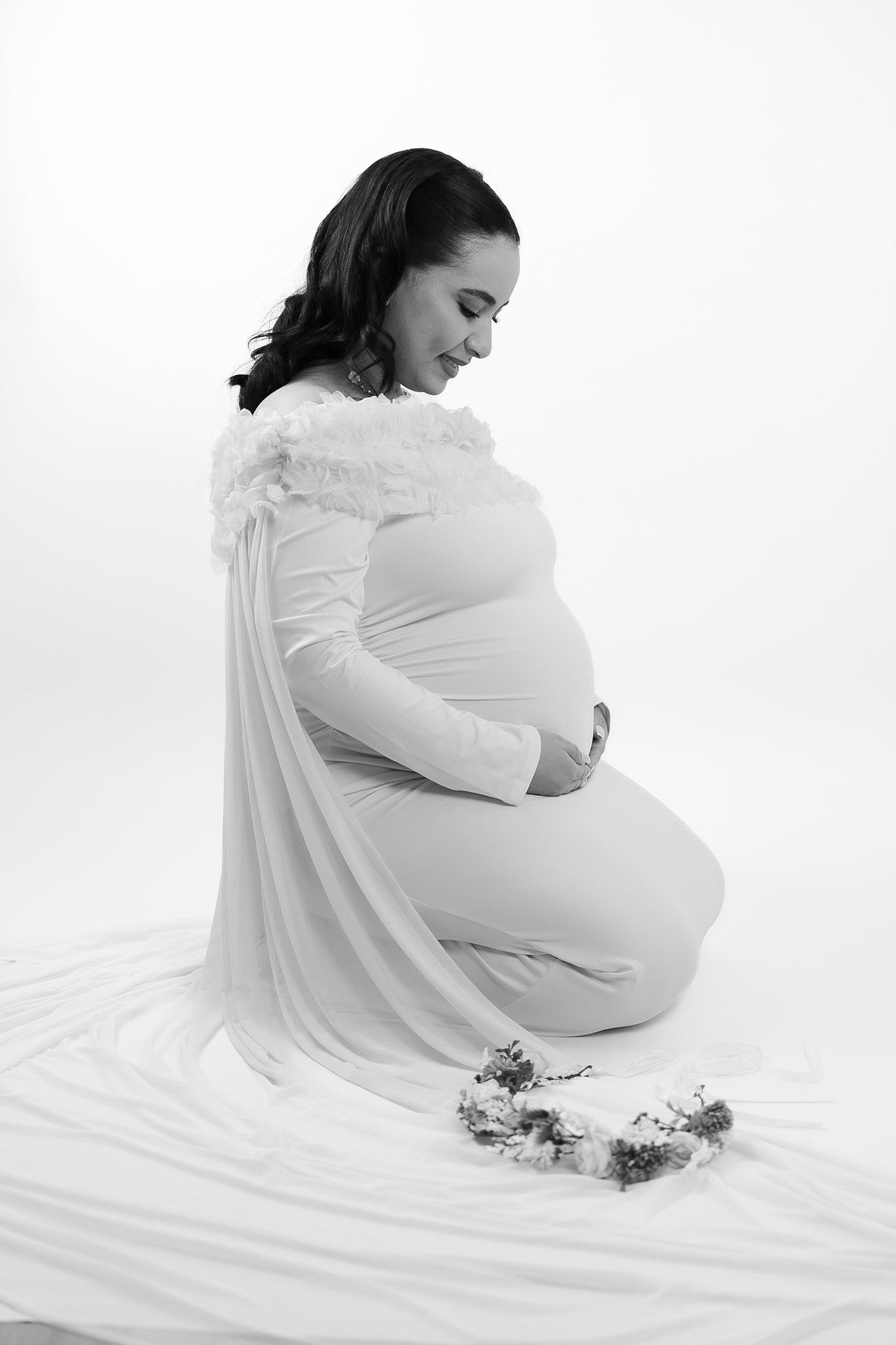 Black and white maternity photo of pregnant woman in white dress kneeling, cradling baby bump with floral crown nearby.