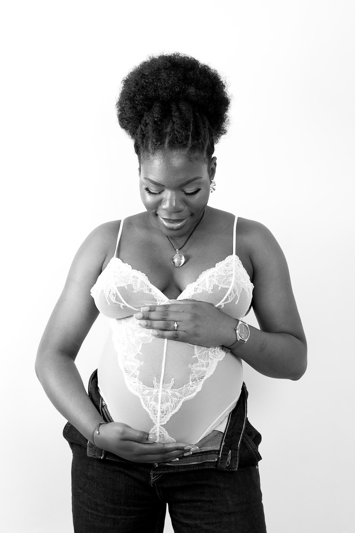 Black and white portrait of a smiling pregnant woman in lace bodysuit and jeans, cradling her baby bump.
