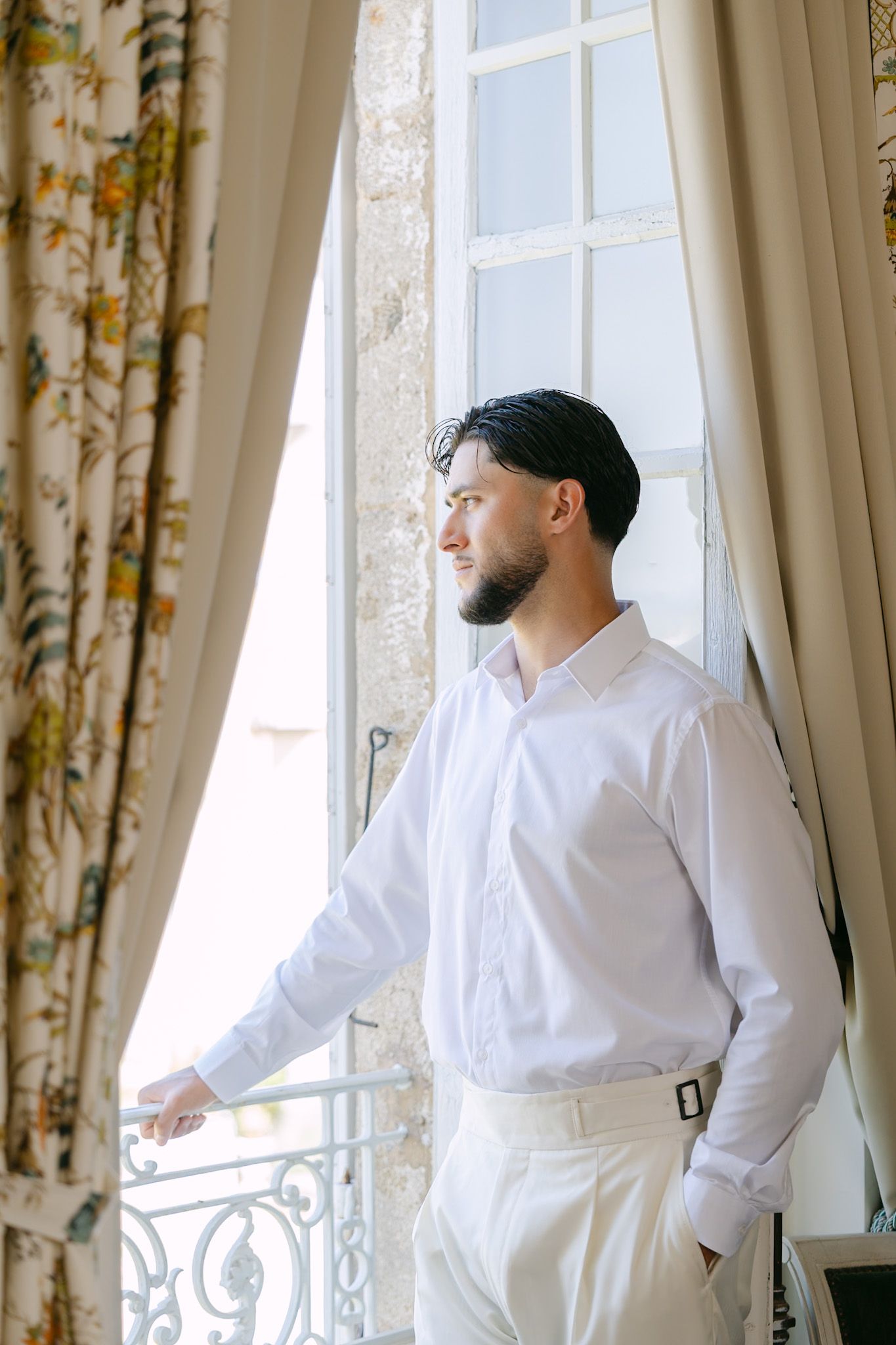 Bearded man in white shirt and pants standing by an elegant window with floral curtains, gazing outside thoughtfully.