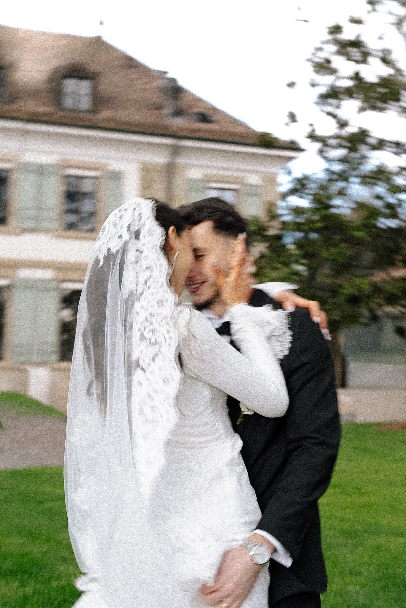 Joyful bride and groom embracing outdoors, bride in lace dress and veil, groom in black suit.