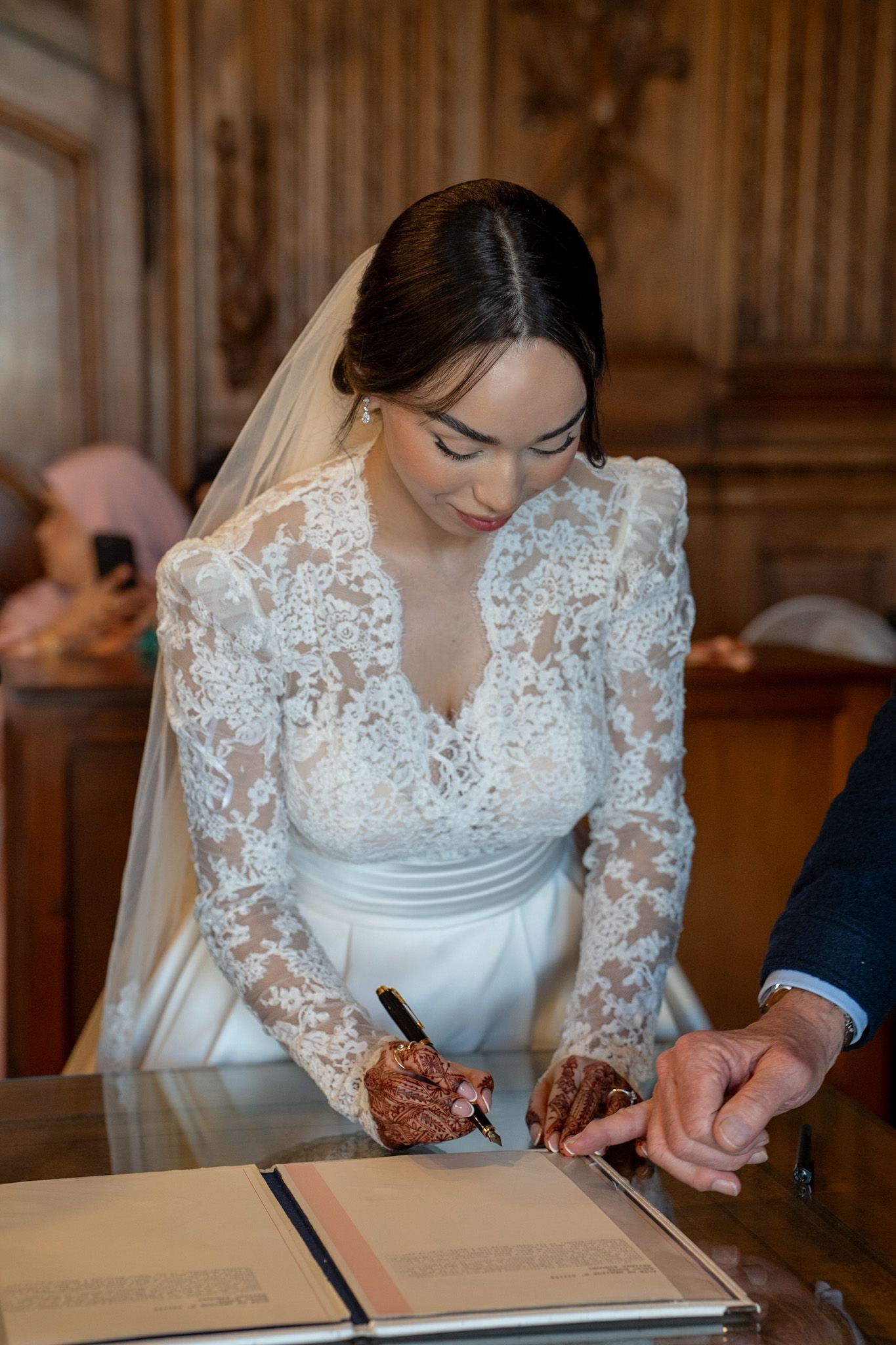 Bride in lace wedding dress signing marriage certificate with henna hands at elegant wooden desk.