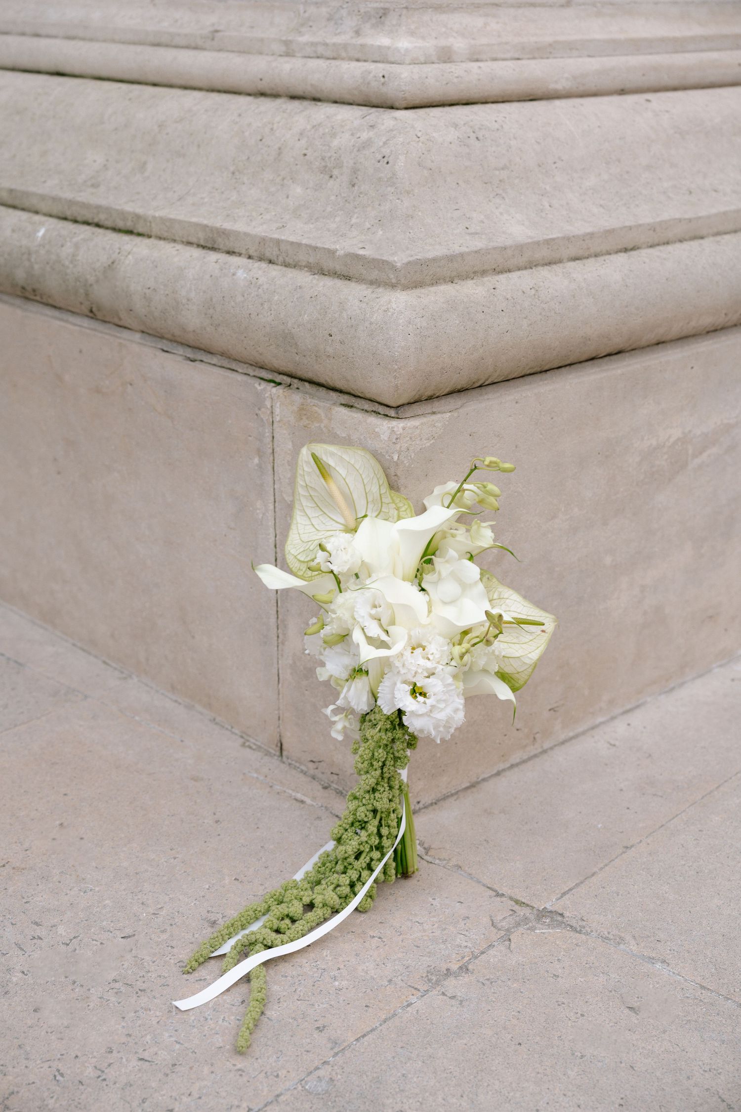 White wedding bouquet with calla lilies and roses leaning against a stone column on a marble floor.