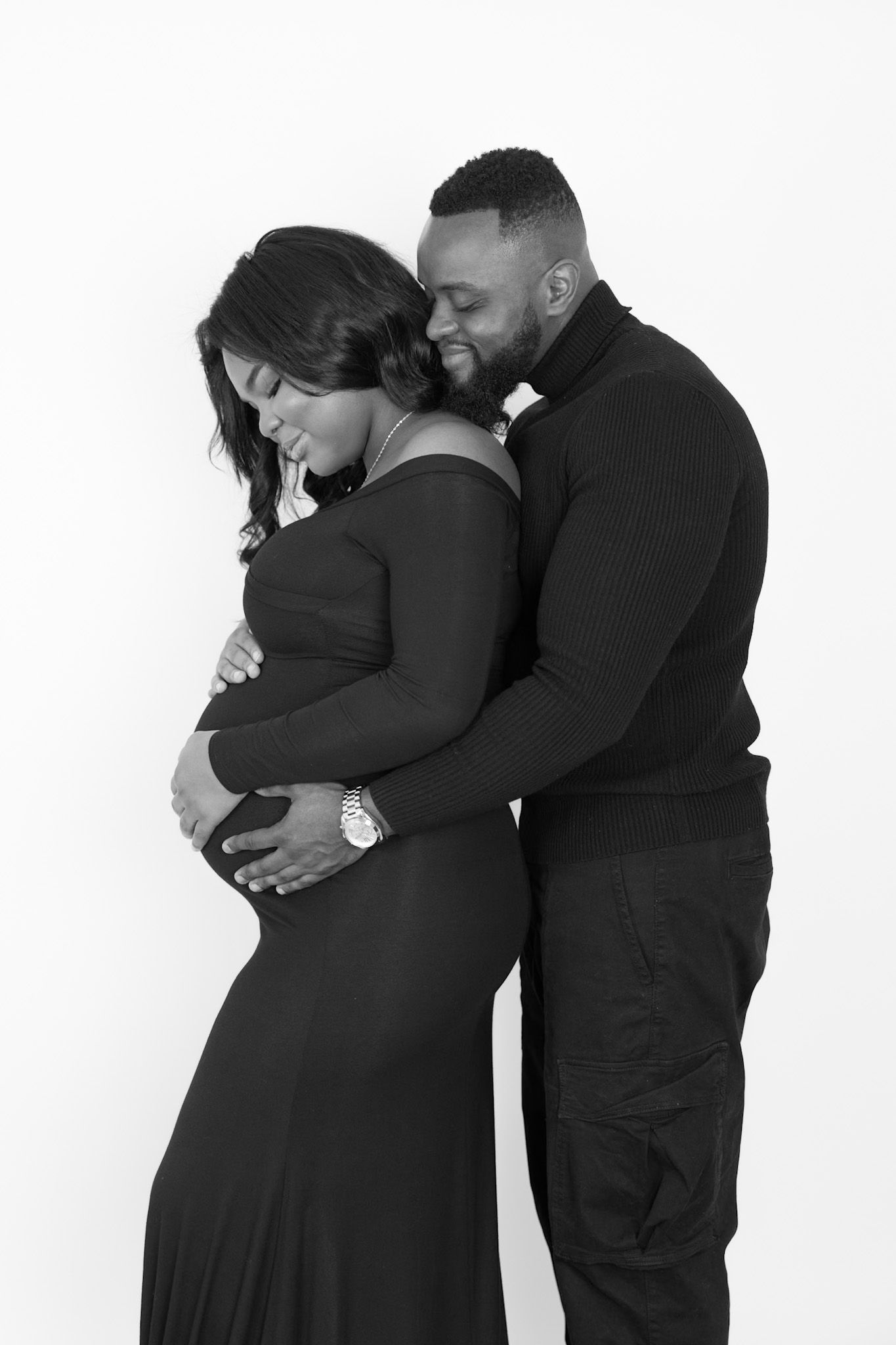 Black and white maternity photo of a couple embracing, man hugging pregnant woman from behind, both smiling tenderly.
