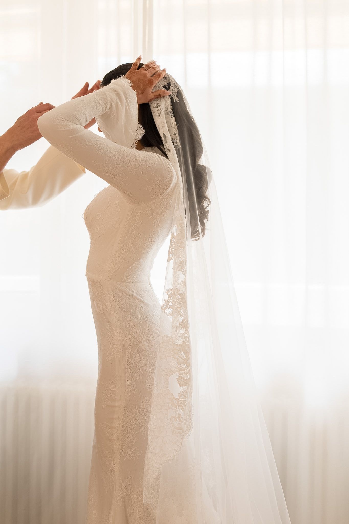 Bride in white lace gown with long veil being adjusted near bright window on wedding day.