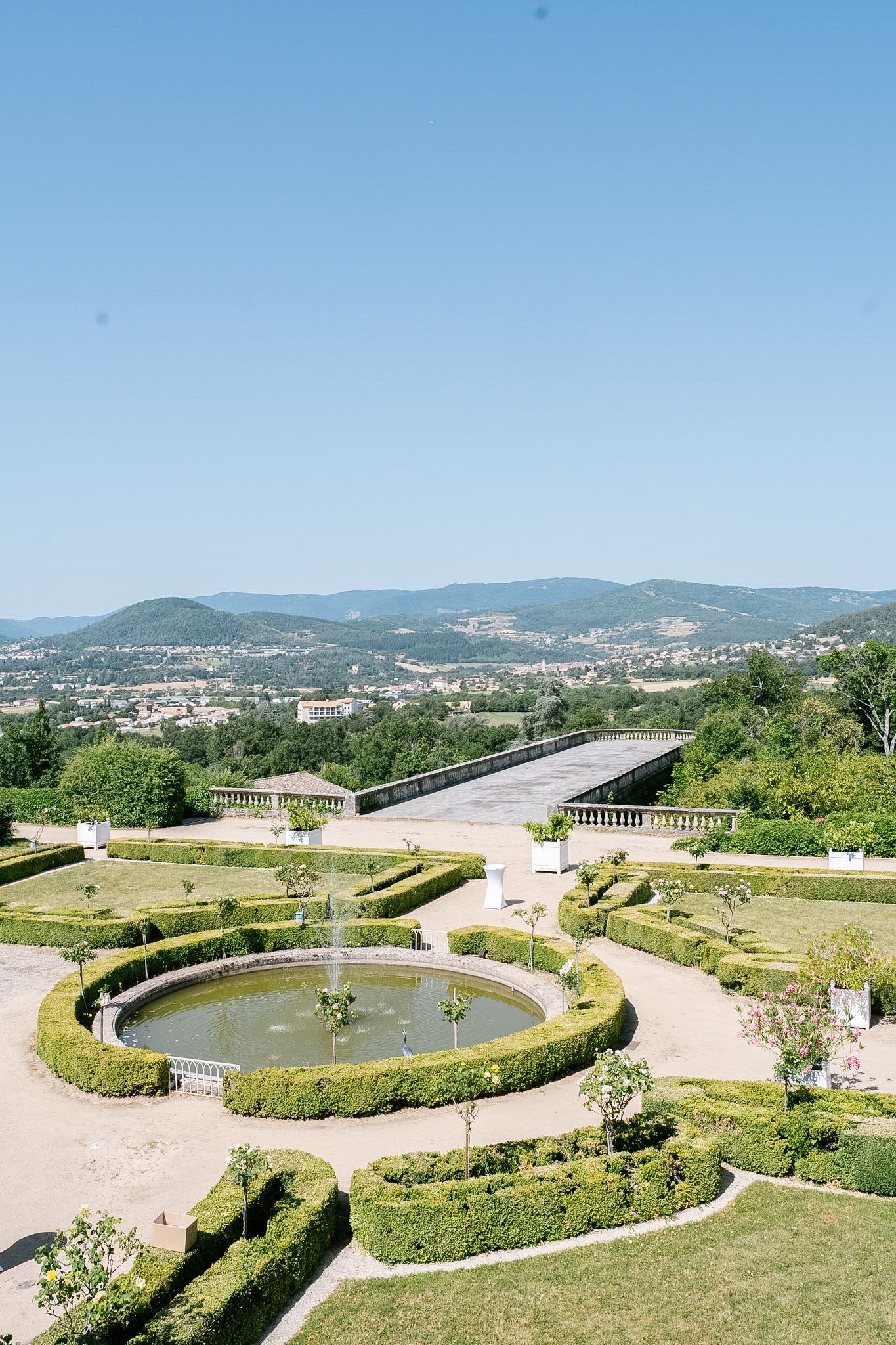 Formal garden with circular fountain, manicured hedges, and panoramic mountain city views under clear blue sky.