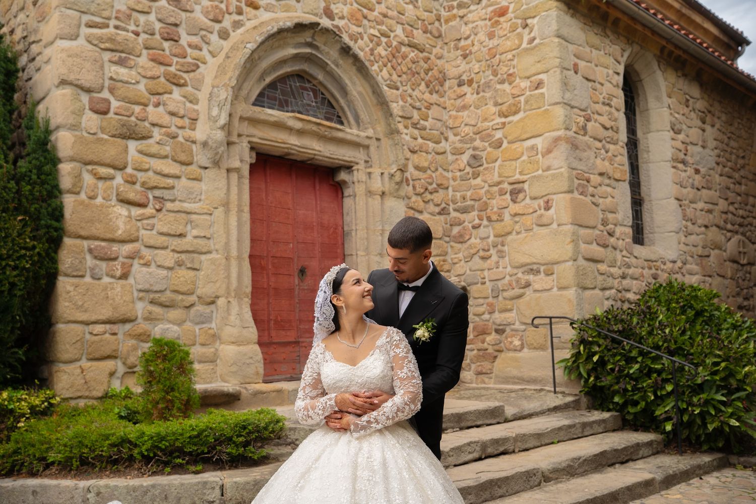 Couple poses in front of rustic stone church wall with red wooden doors and arched architectural details.
