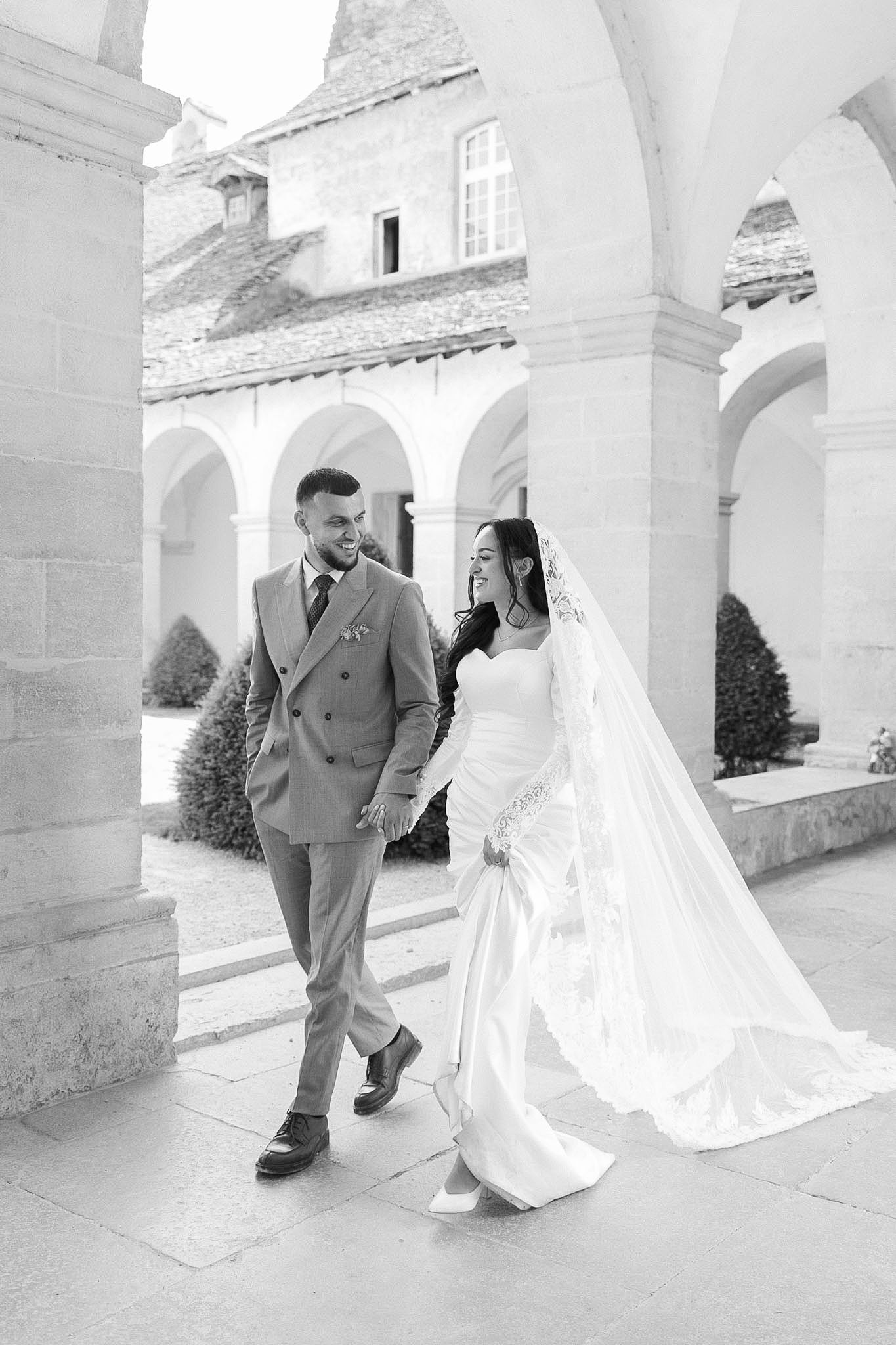 Black and white photo of a bride and groom walking hand in hand through elegant stone arched courtyard.