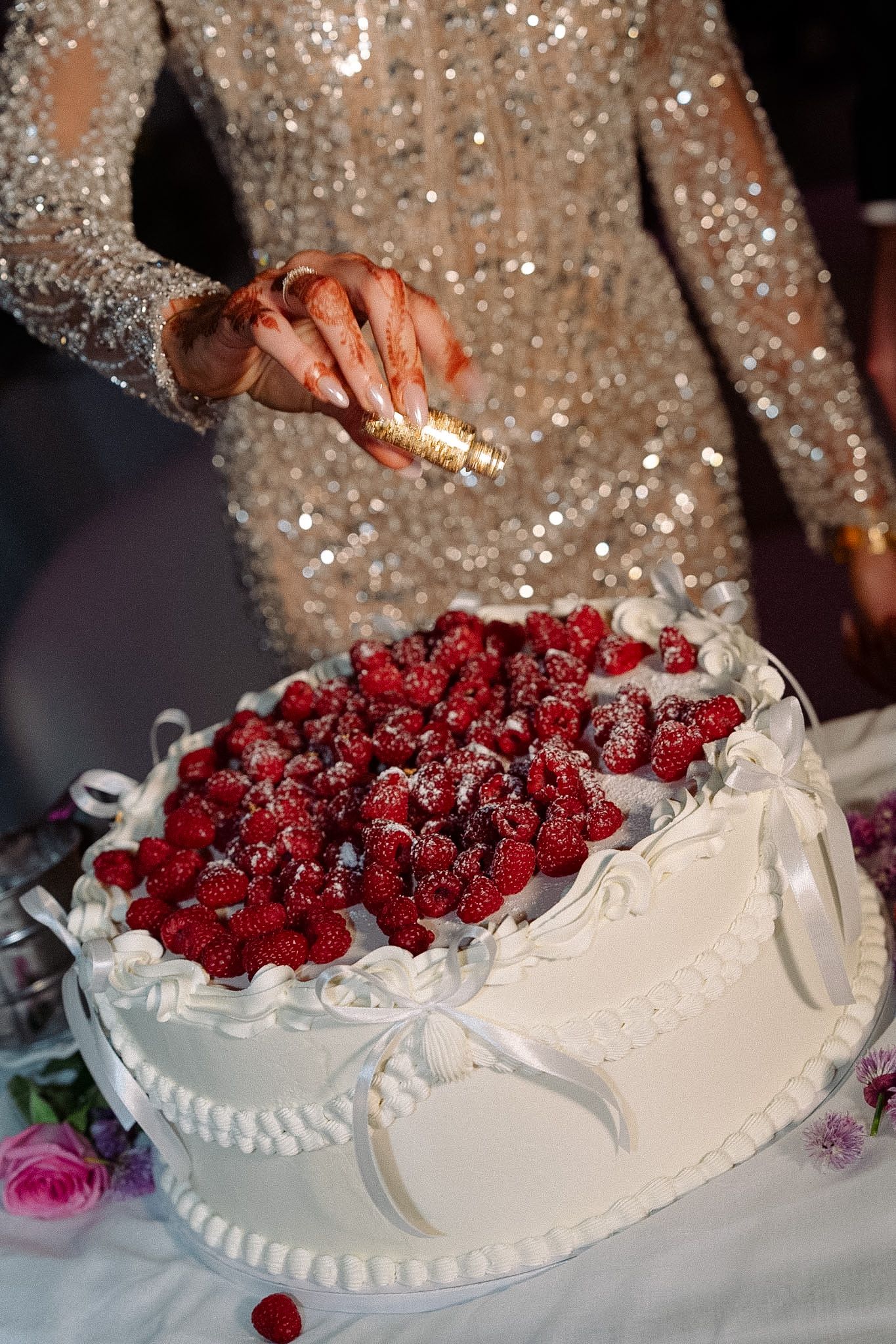 Person in sequined dress sprinkling sugar on a white wedding cake topped with fresh raspberries.