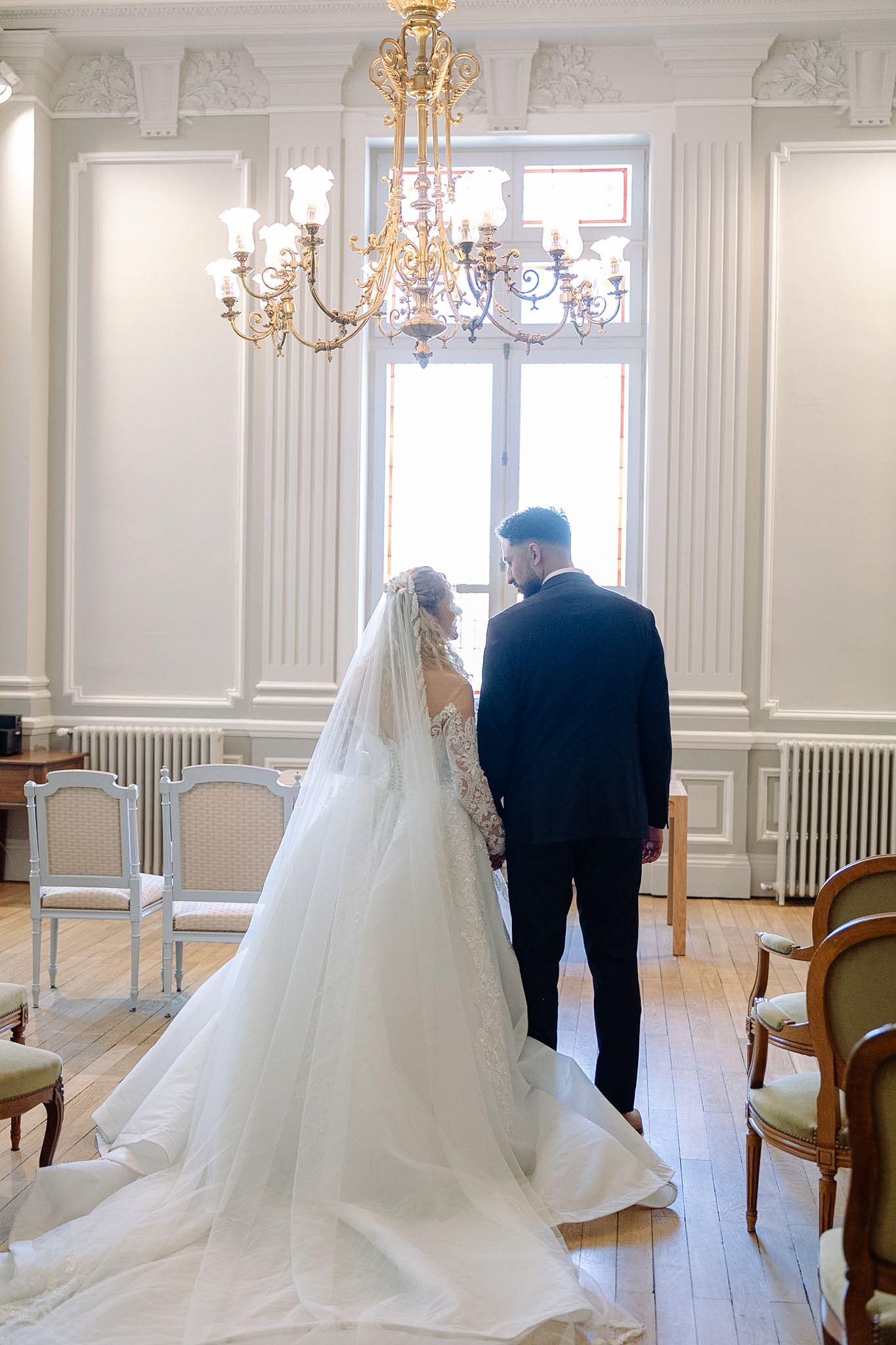 Bride and groom standing hand in hand before a window in an elegant white room with chandelier.
