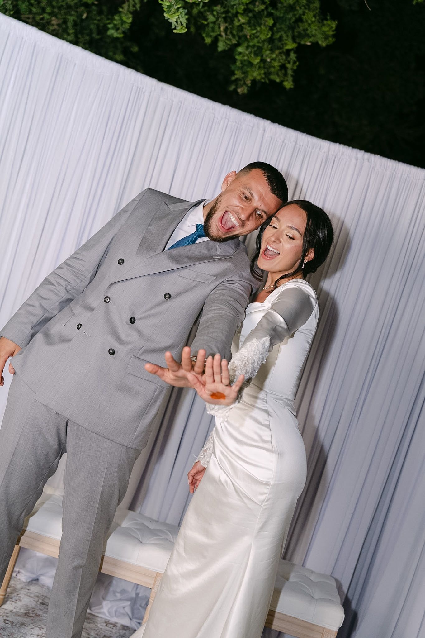 Joyful newlyweds posing together, groom in gray suit and bride in white gown, showing off wedding rings outdoors.