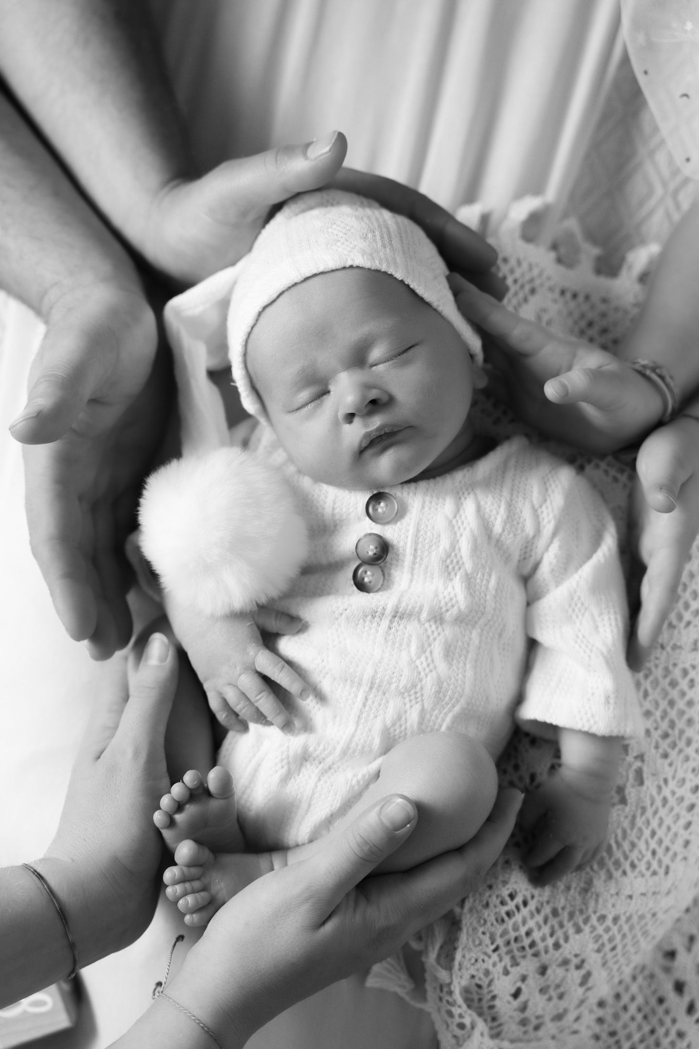 Black and white photo of a sleeping newborn baby in a knit outfit, cradled gently by parents hands.