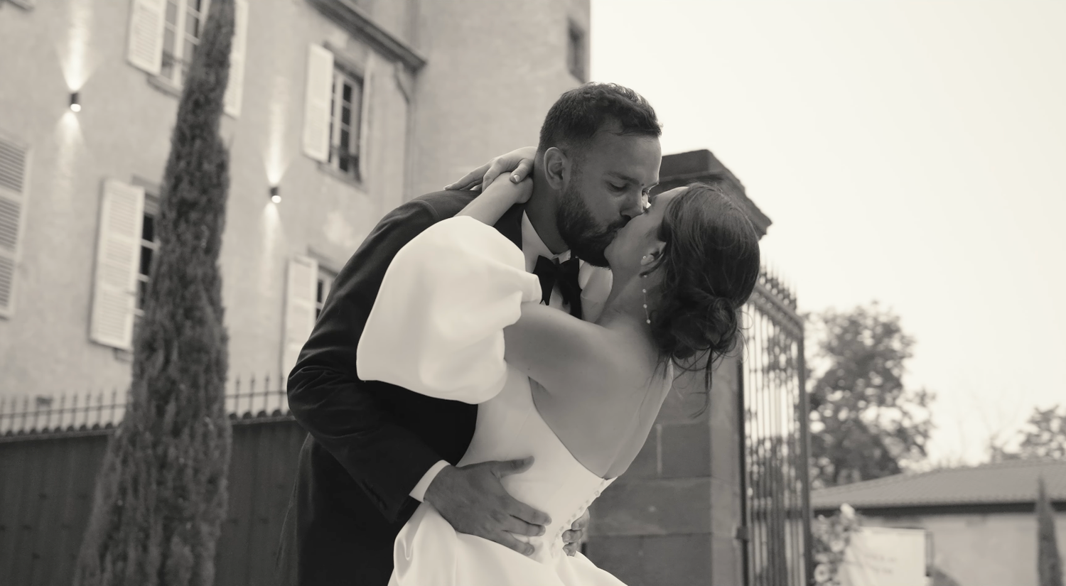 Black and white photo of a bride and groom sharing a romantic kiss outdoors near a historic building.