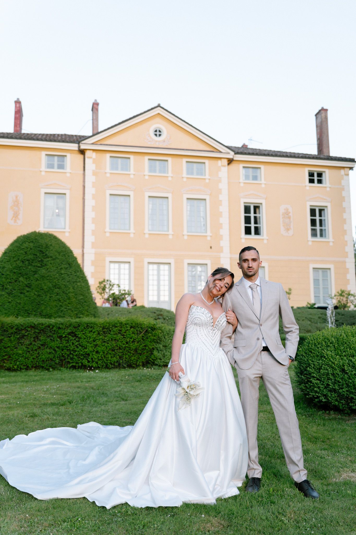 Bride and groom posing in front of elegant yellow French chateau surrounded by manicured hedges on their wedding day.