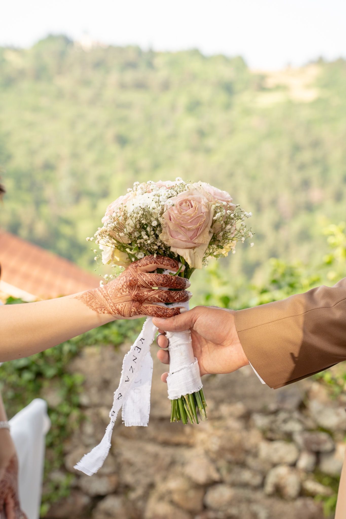 Two hands exchange a delicate pink rose and baby's breath bouquet with white ribbon against a blurred outdoor background.