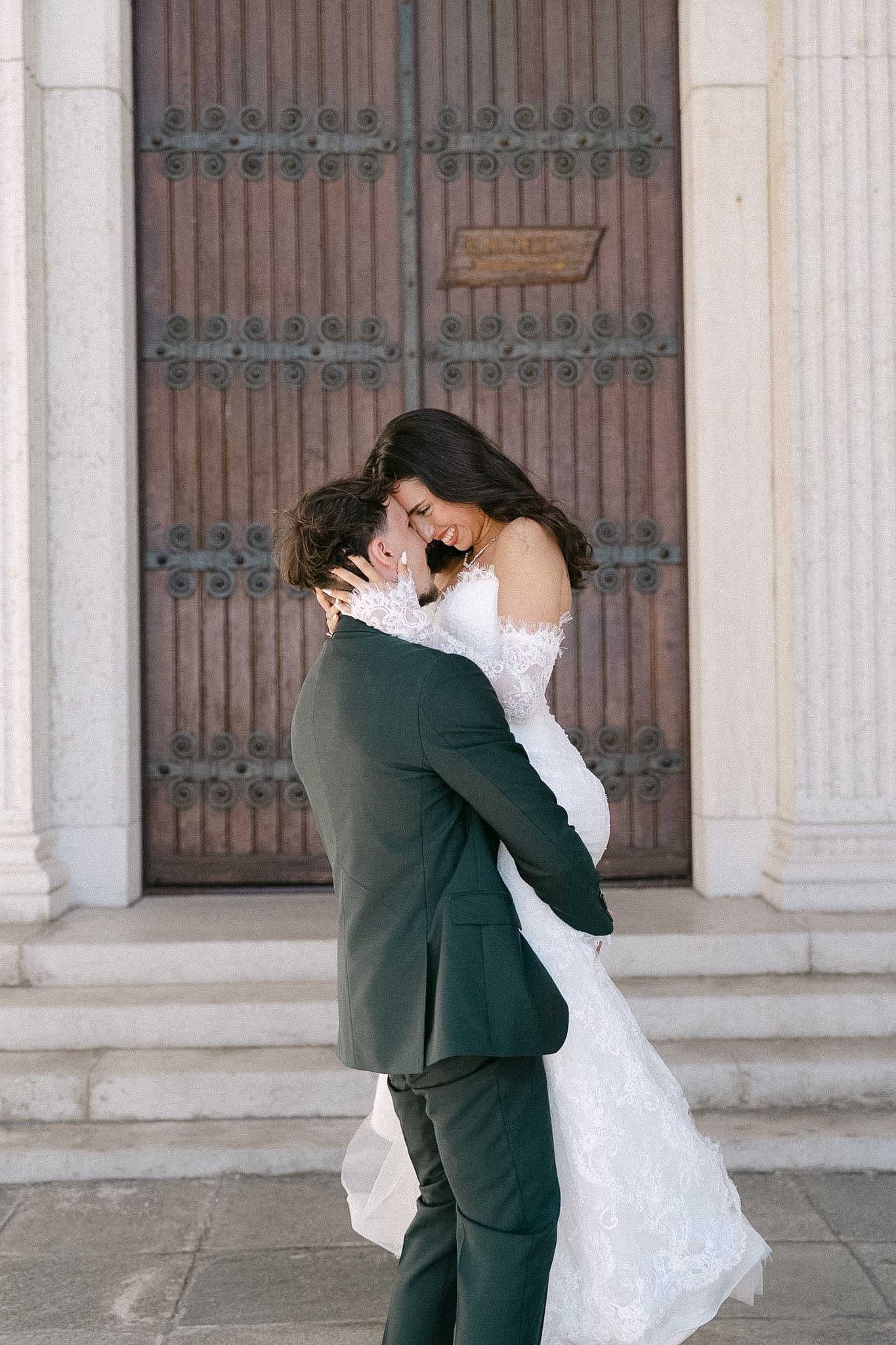 Bride and groom embracing joyfully before grand wooden doors, she in lace off-shoulder gown, he in dark green suit.