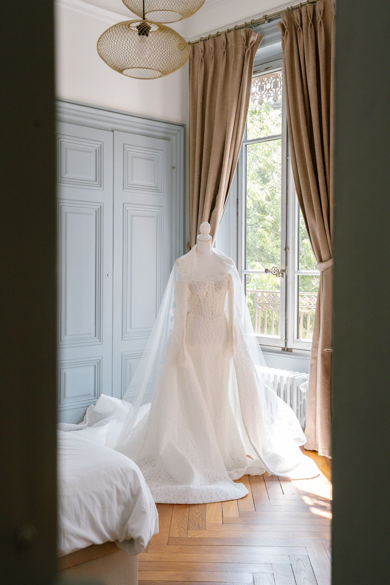 Elegant white wedding gown with flowing veil displayed on mannequin in sunlit French chateau bedroom with herringbone floors.