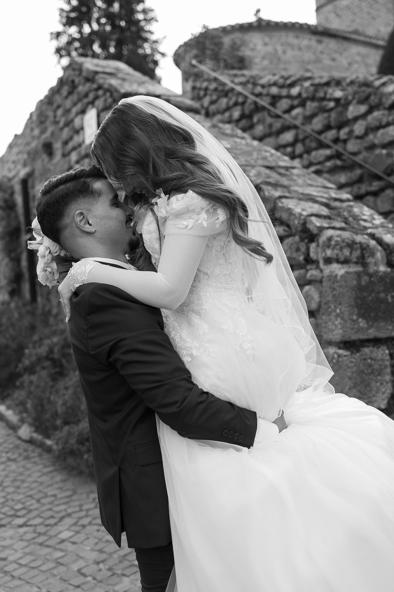Black and white photo of a bride and groom sharing a romantic kiss outdoors near a stone building.