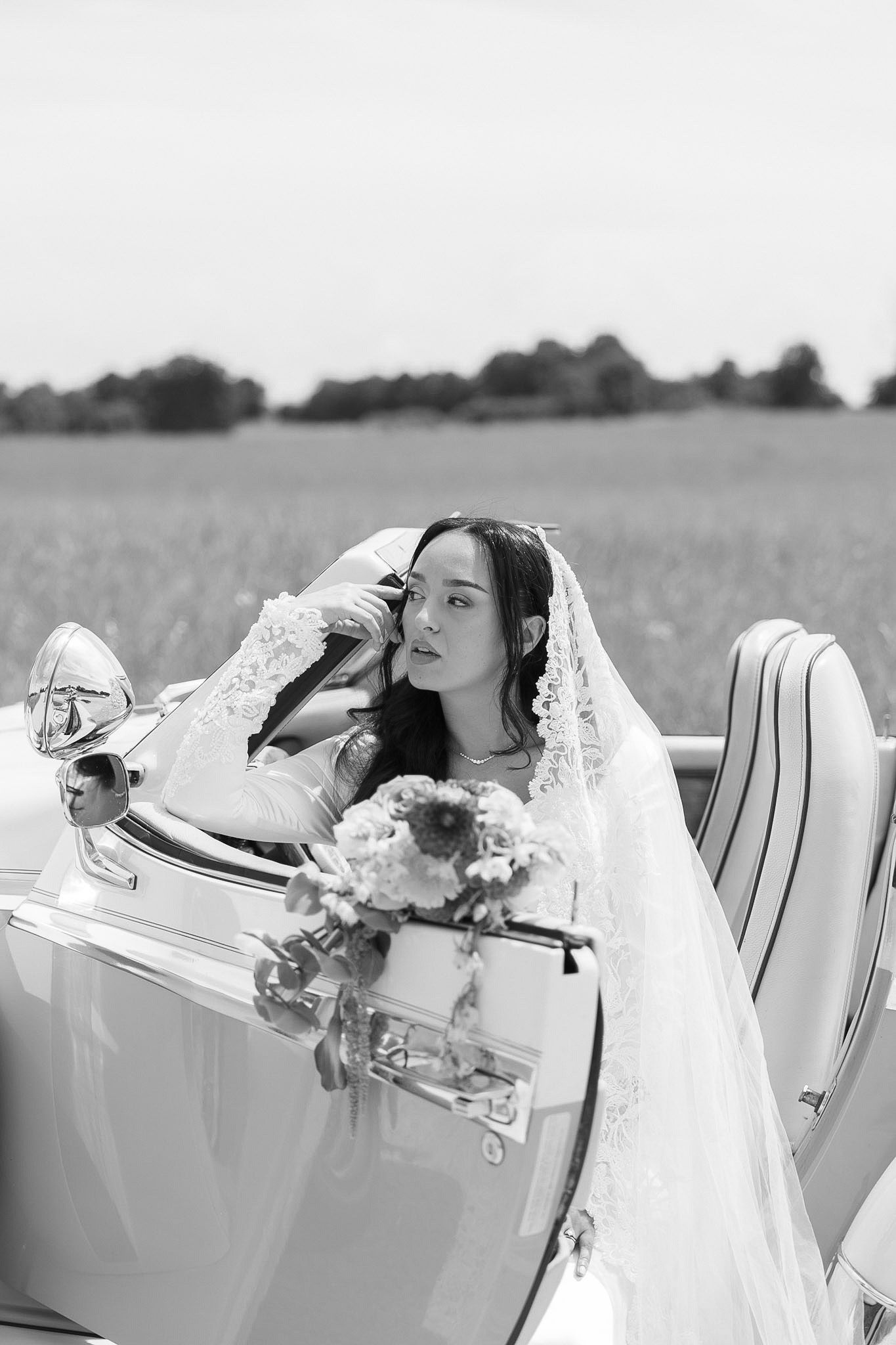 Black and white photo of a bride in veil sitting in a vintage convertible car with a bouquet, field in background.