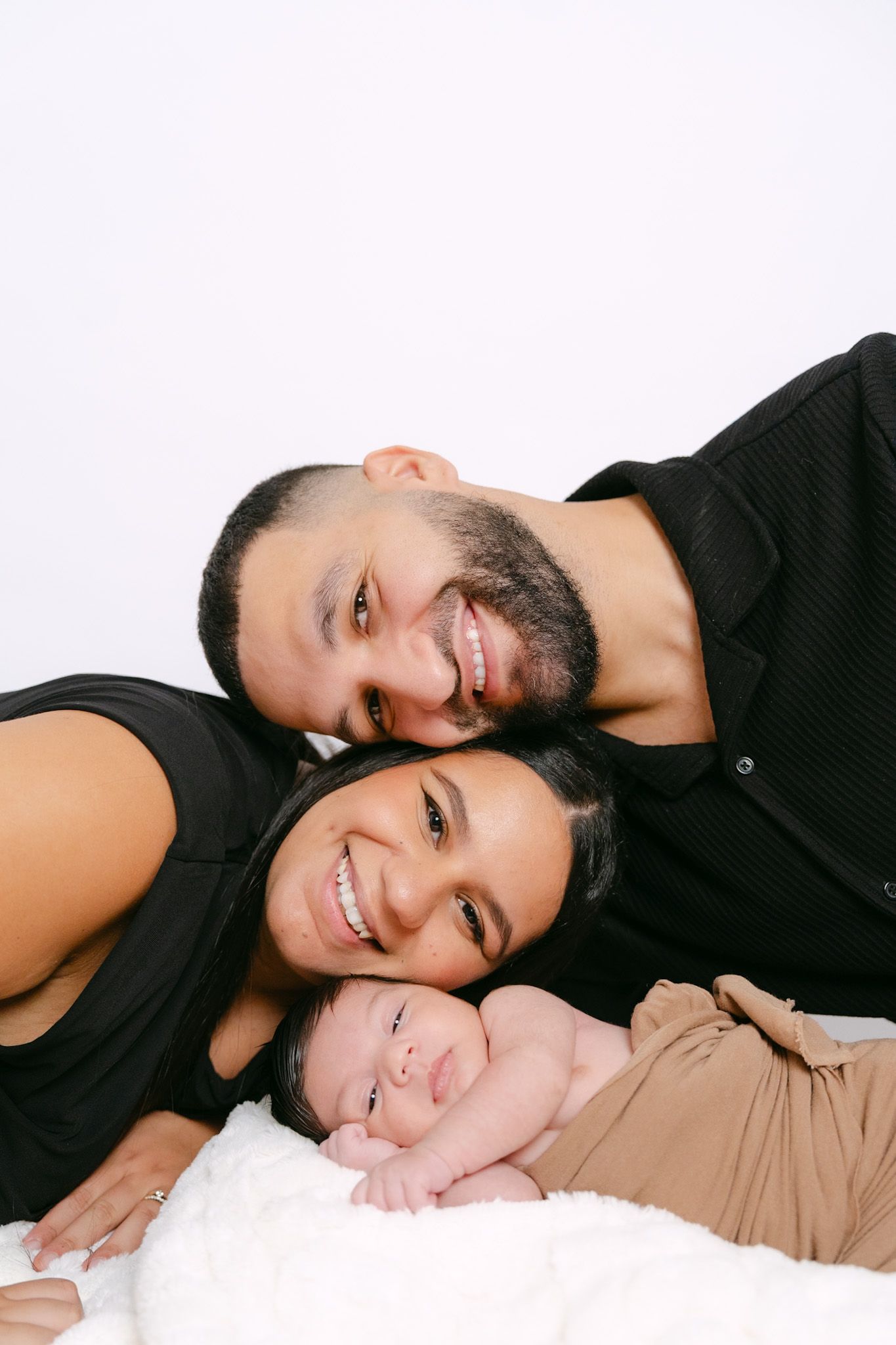 Happy couple in black outfits lying down smiling with their newborn baby on a white surface.