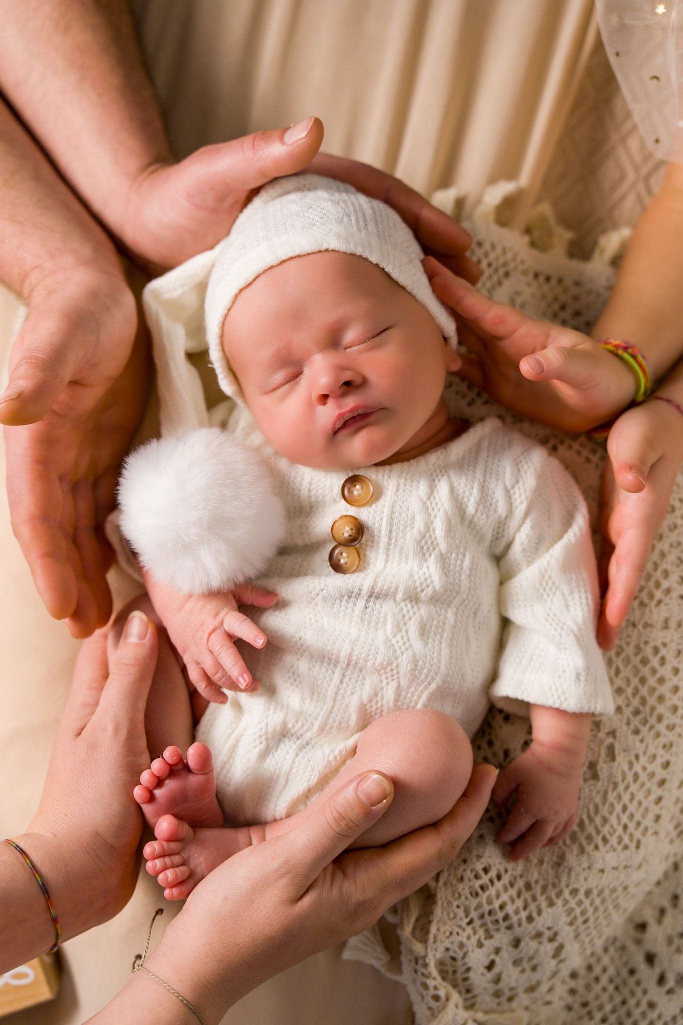 A newborn baby wearing a white knit outfit and cap is gently cradled in caring hands on a lace blanket.