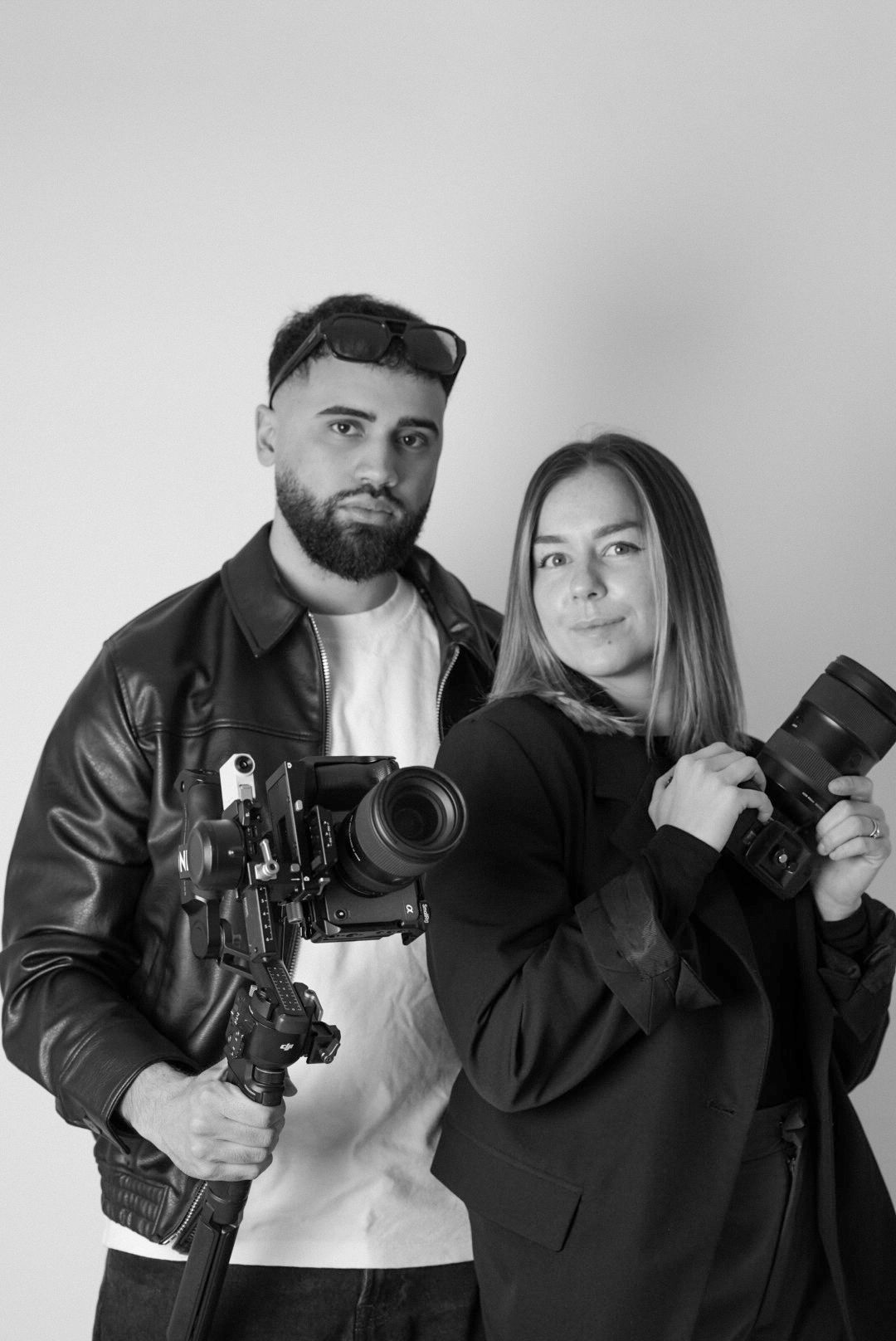 Black and white photo of two photographers, man holding video camera rig and woman holding DSLR camera.