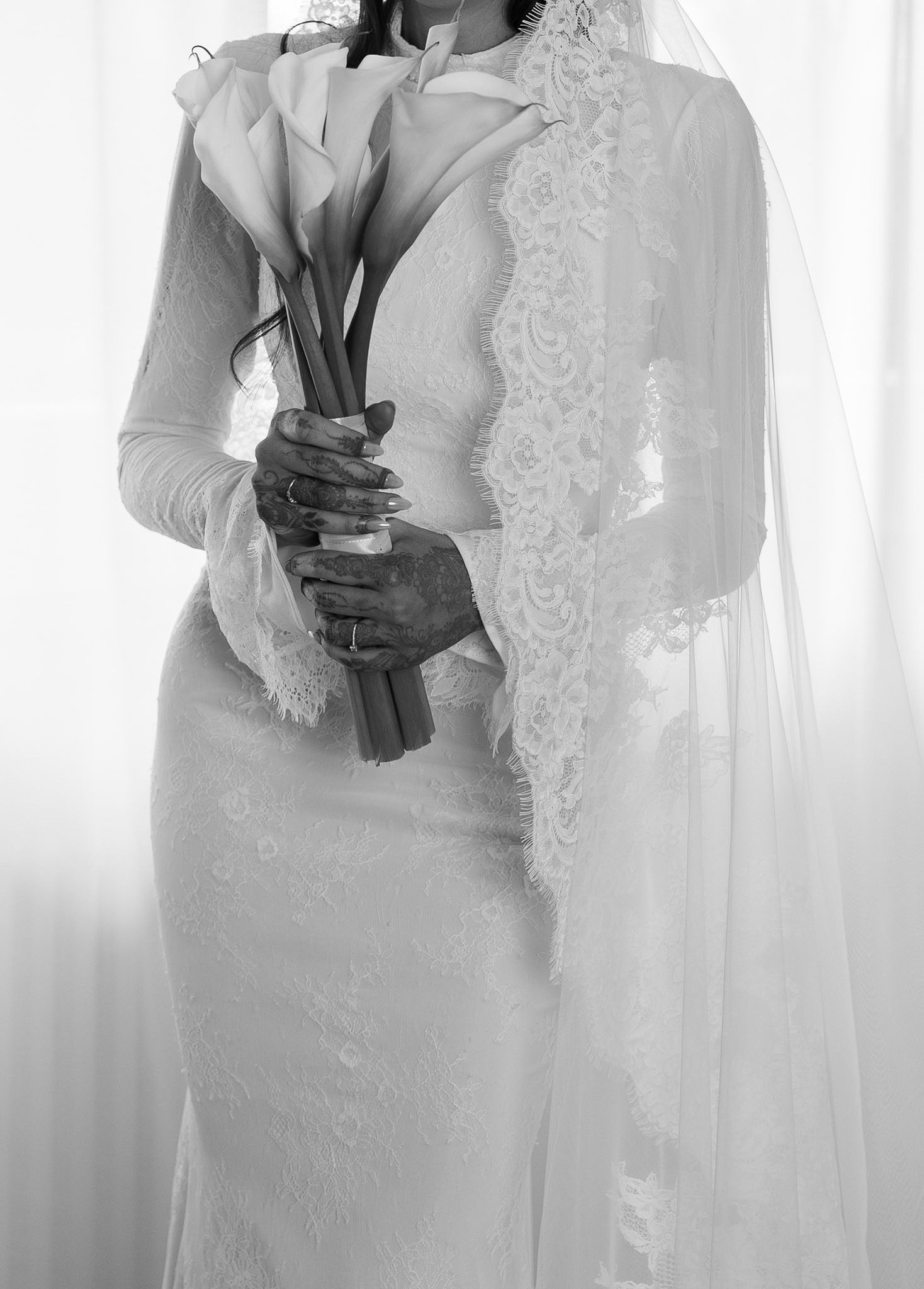 Black and white photo of a bride in a lace wedding gown and veil holding a calla lily bouquet.