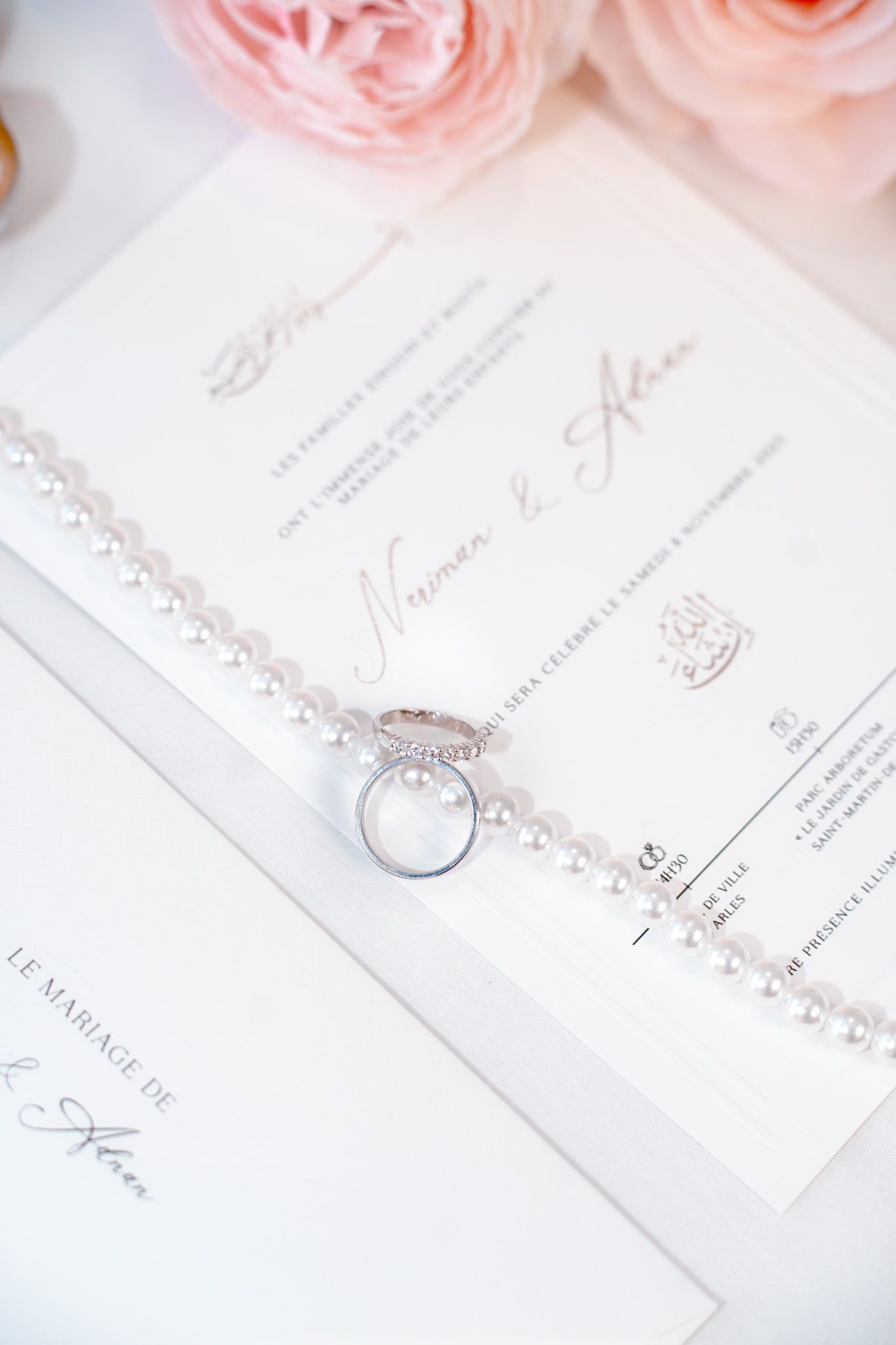 Elegant wedding flat lay with diamond ring on pearl necklace, French invitation cards, and pink roses on white background.