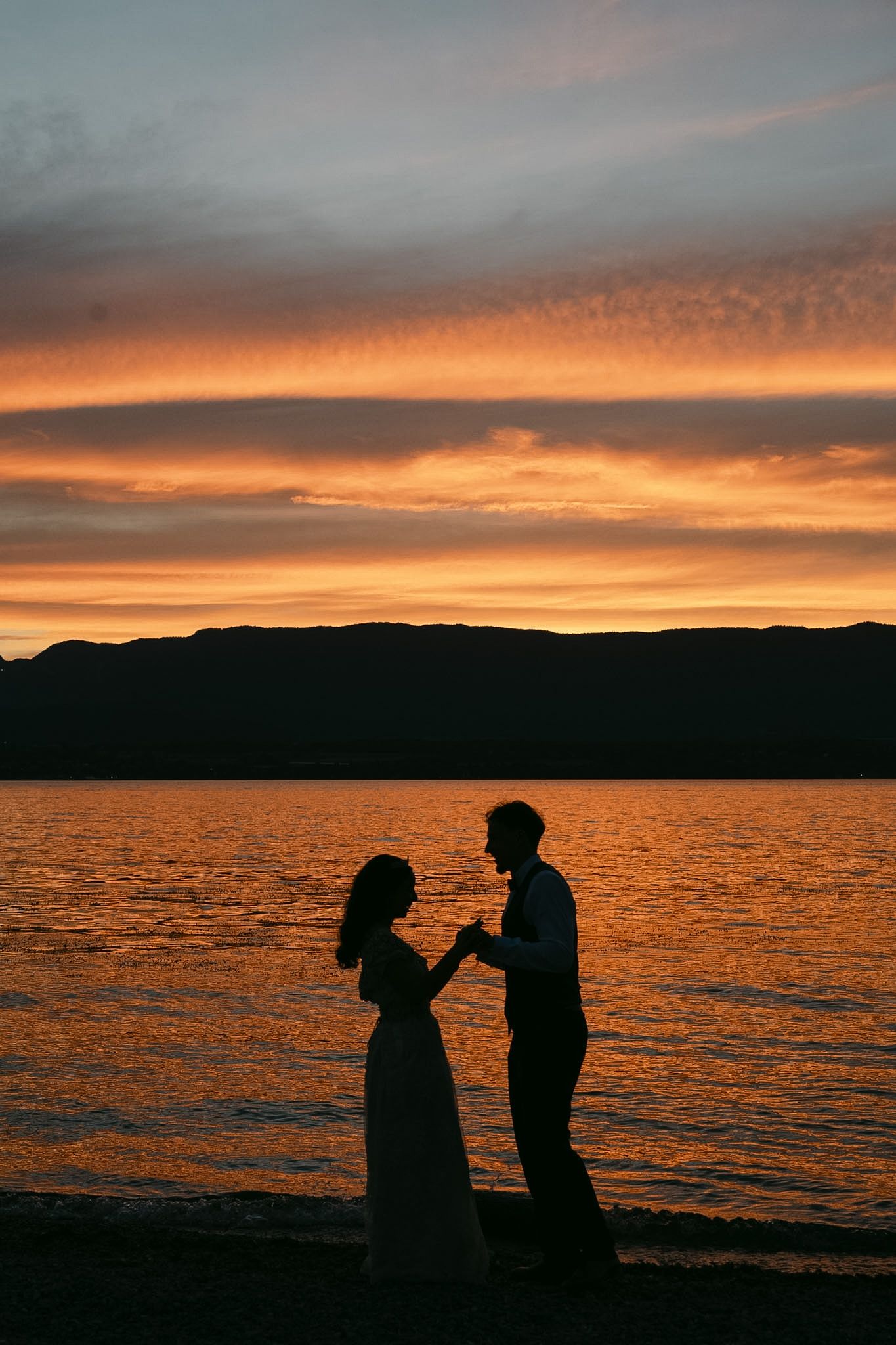 Silhouette of a couple holding hands by a lake at sunset with vibrant orange sky and mountain backdrop.