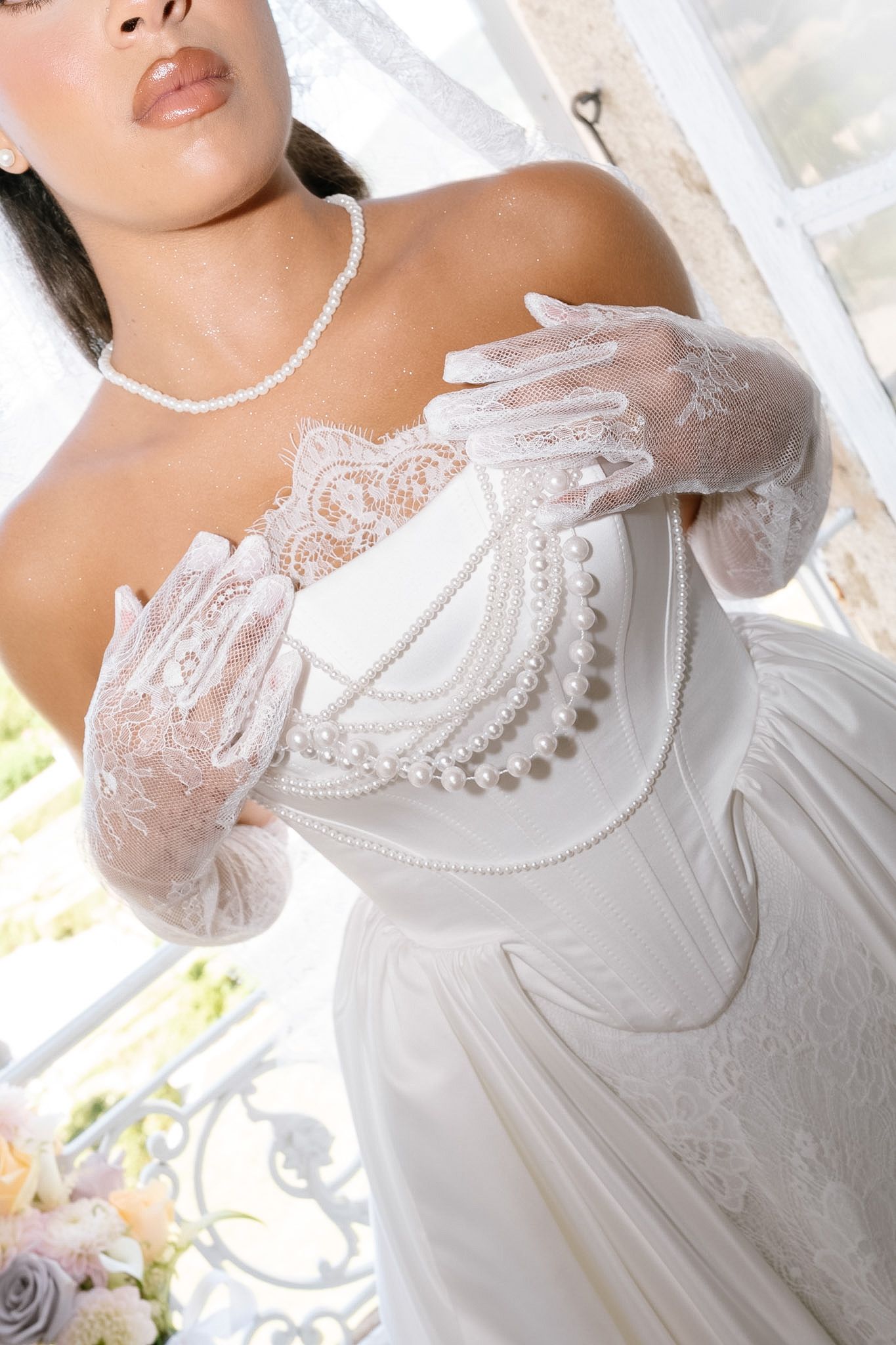 Bride in white off-shoulder lace wedding gown with pearl accessories and lace gloves holding a pearl clutch purse.