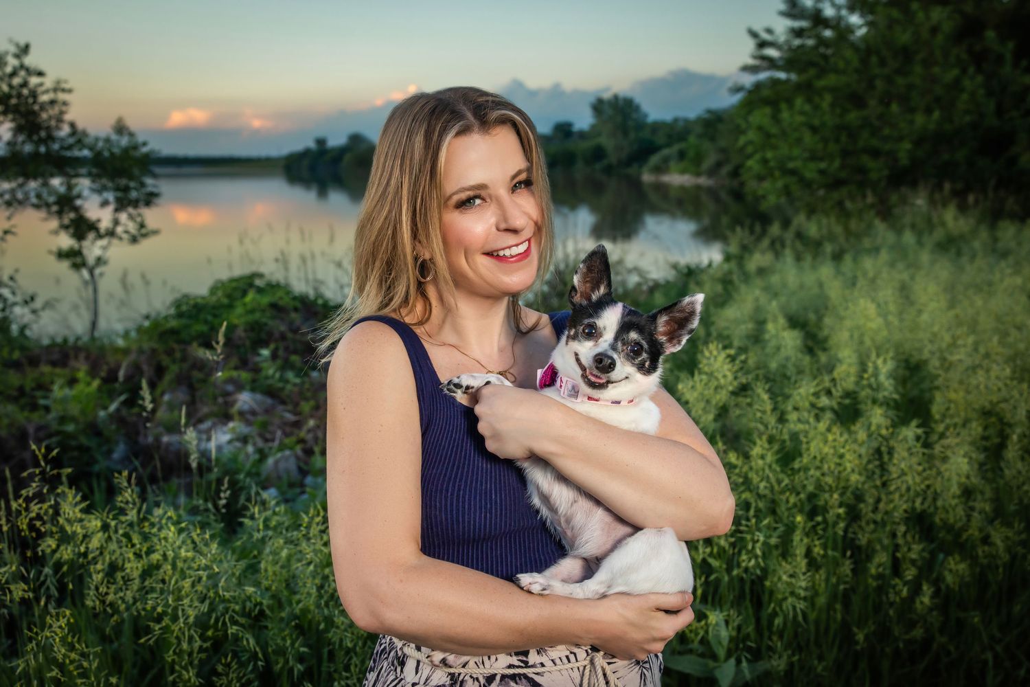 Smiling woman with blonde hair holding small dog near lake at sunset.