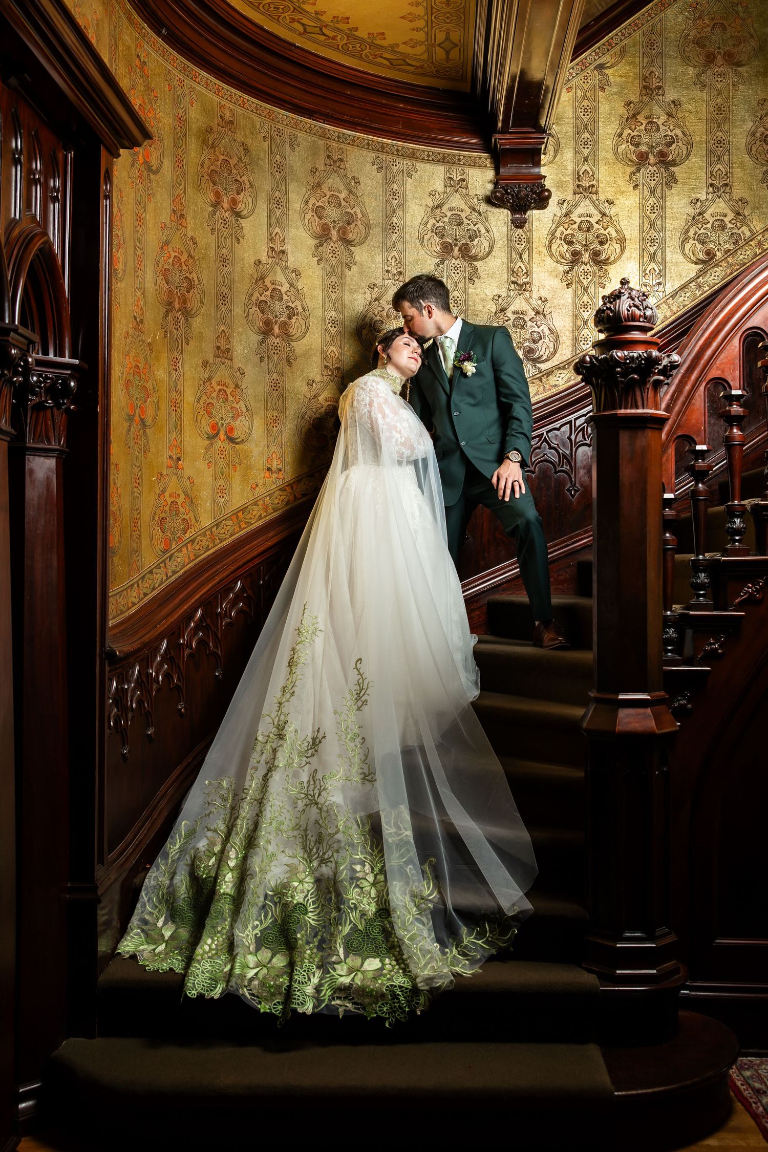 Bride in green floral gown and groom in dark suit share romantic moment on ornate wooden staircase.