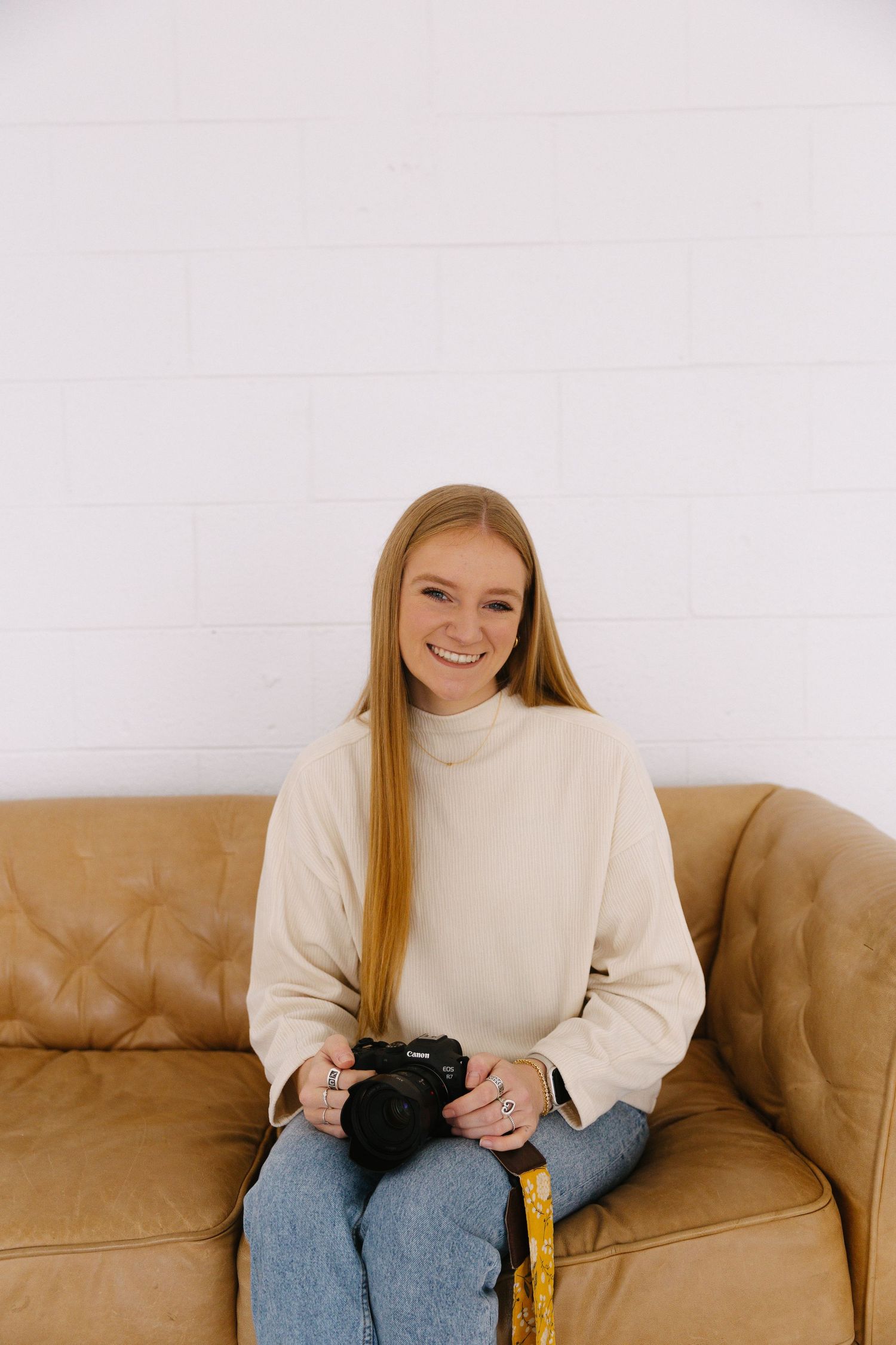 Person in cream turtleneck sweater and jeans sits casually on brown leather couch holding camera.