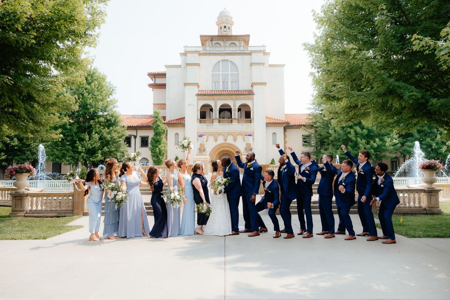 Wedding party in navy and light blue attire posing in front of elegant white mansion.