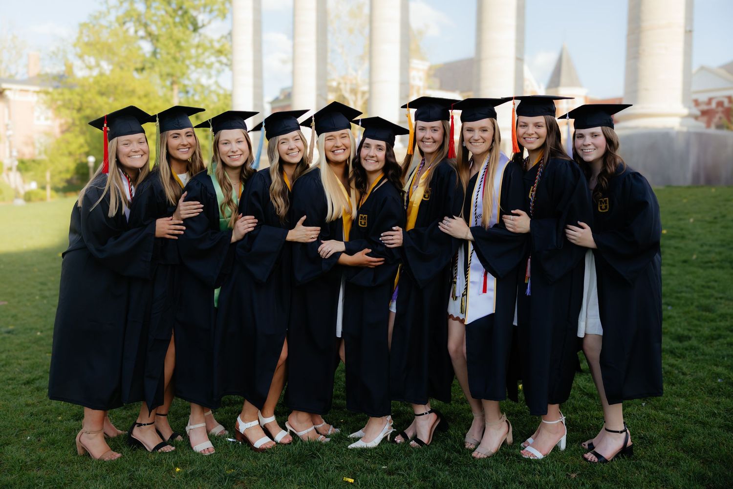 Group of graduates in black caps and gowns pose together on grass for graduation photos.