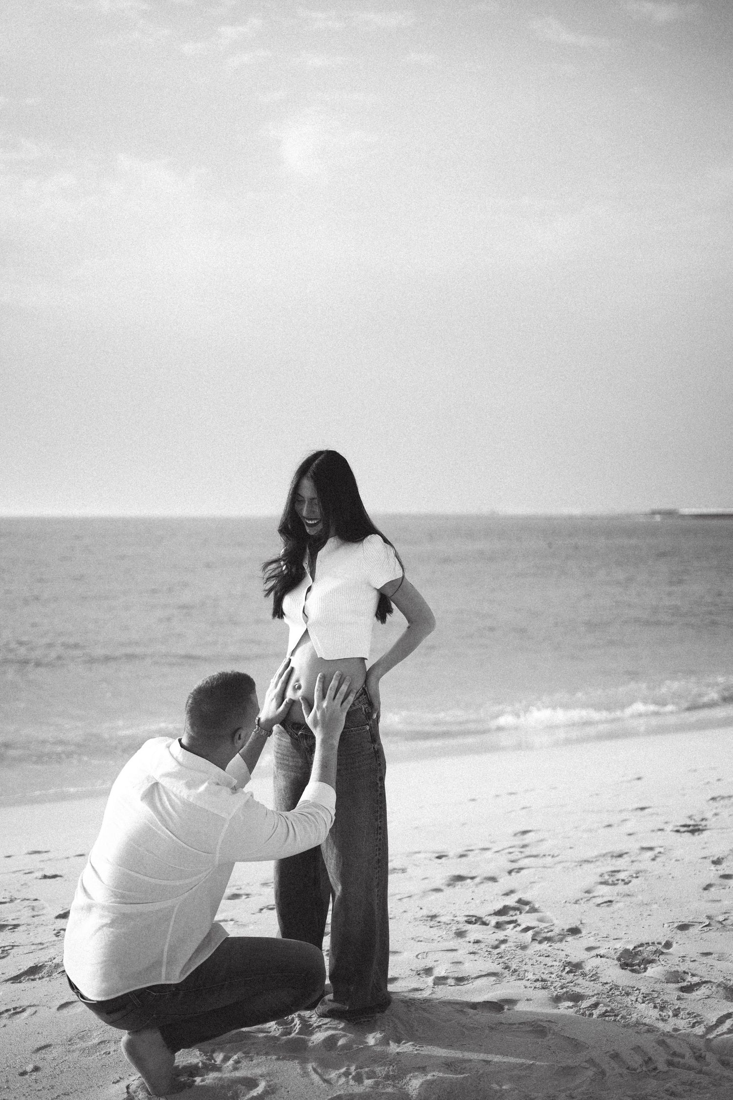 Black and white beach photo of a man kneeling before a pregnant woman, gently touching her baby bump by the ocean.