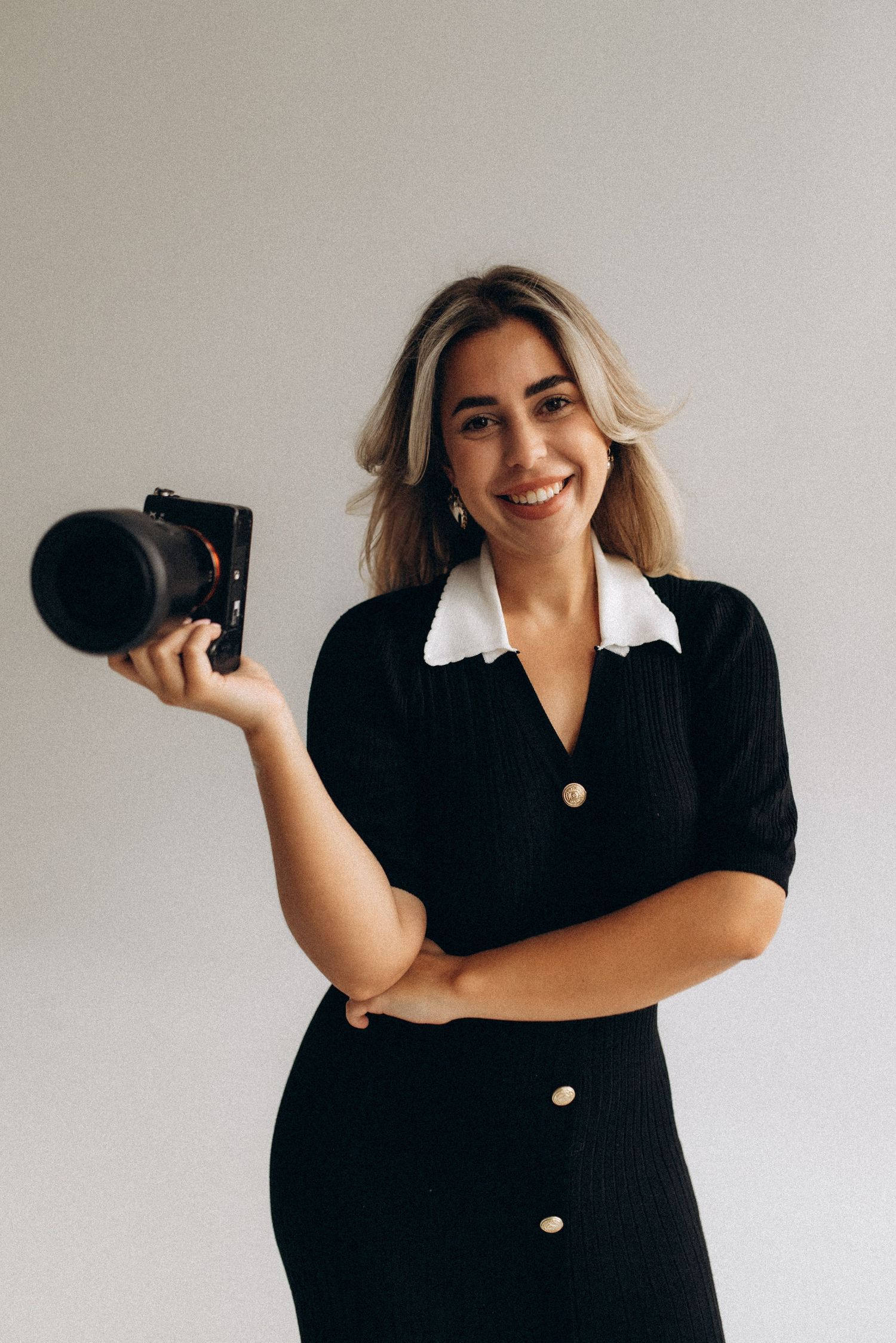 Photographer showing camera while wearing black dress in minimalist studio setup.