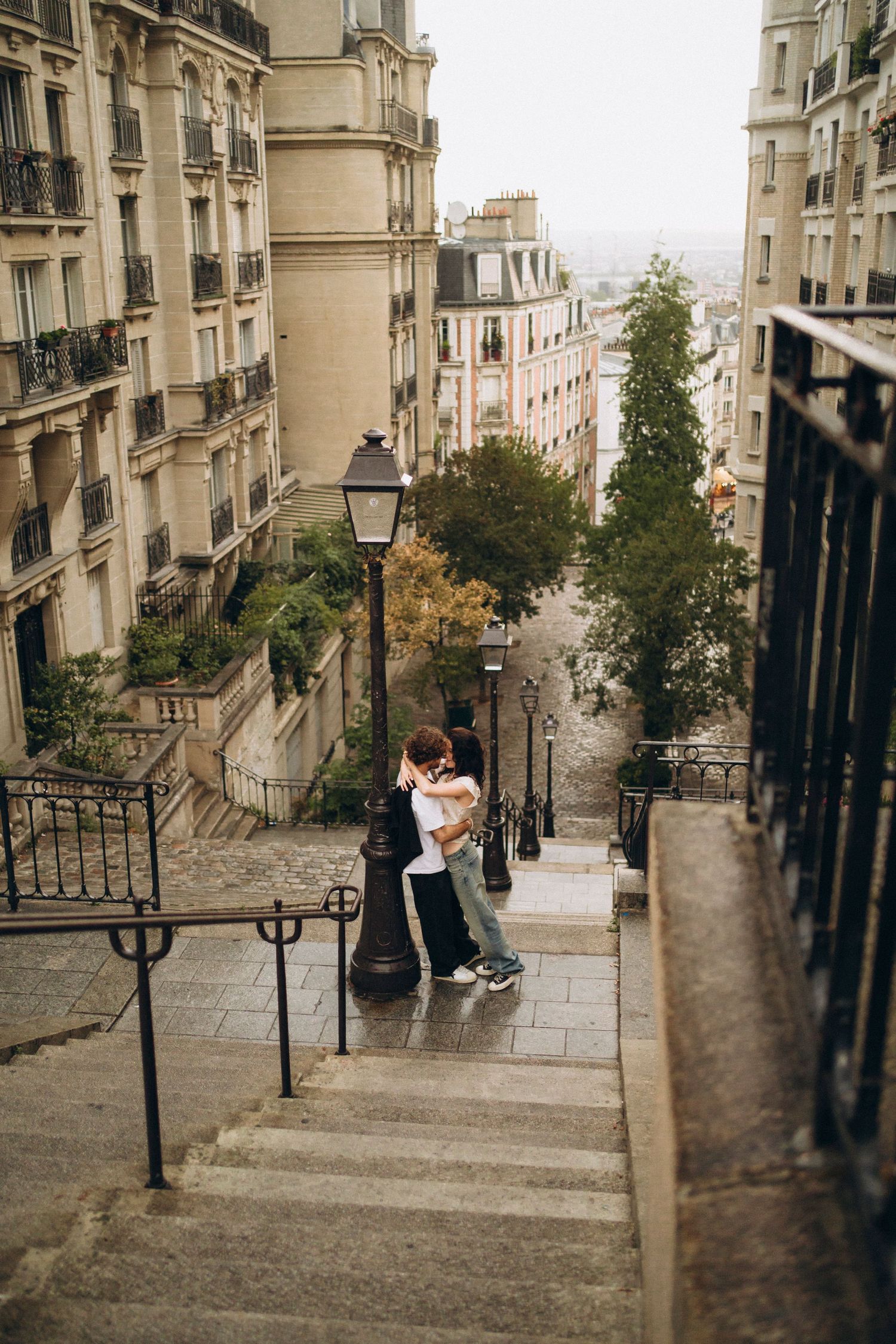 Couple embracing on the iconic steps of Montmartre in Paris, surrounded by classic Haussmann buildings on a cloudy day.