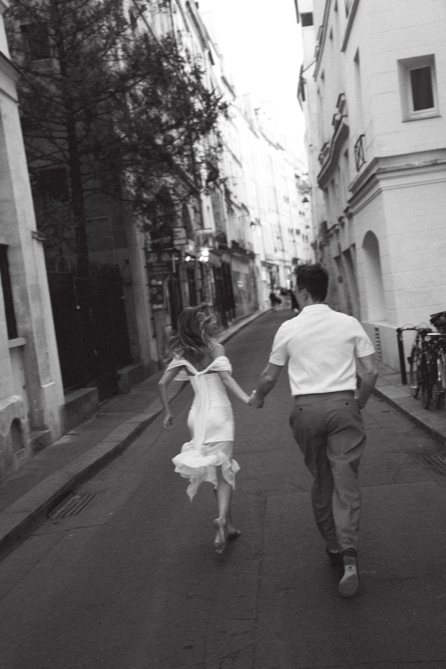Black and white photo of a couple holding hands running down a charming European cobblestone street.