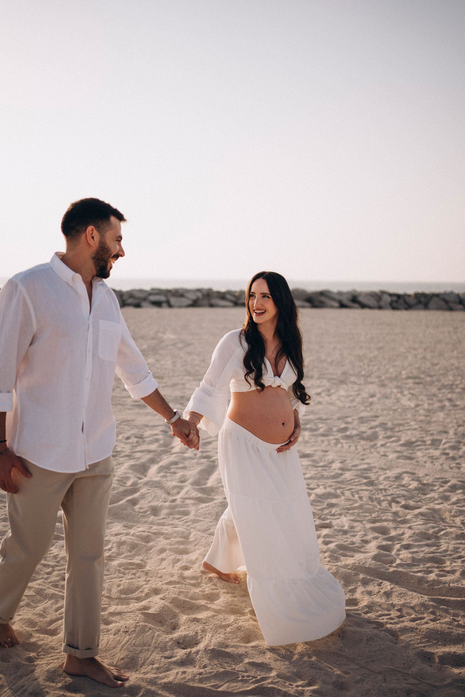 Couple walking and laughing on beach, woman in flowing white dress showing baby bump.