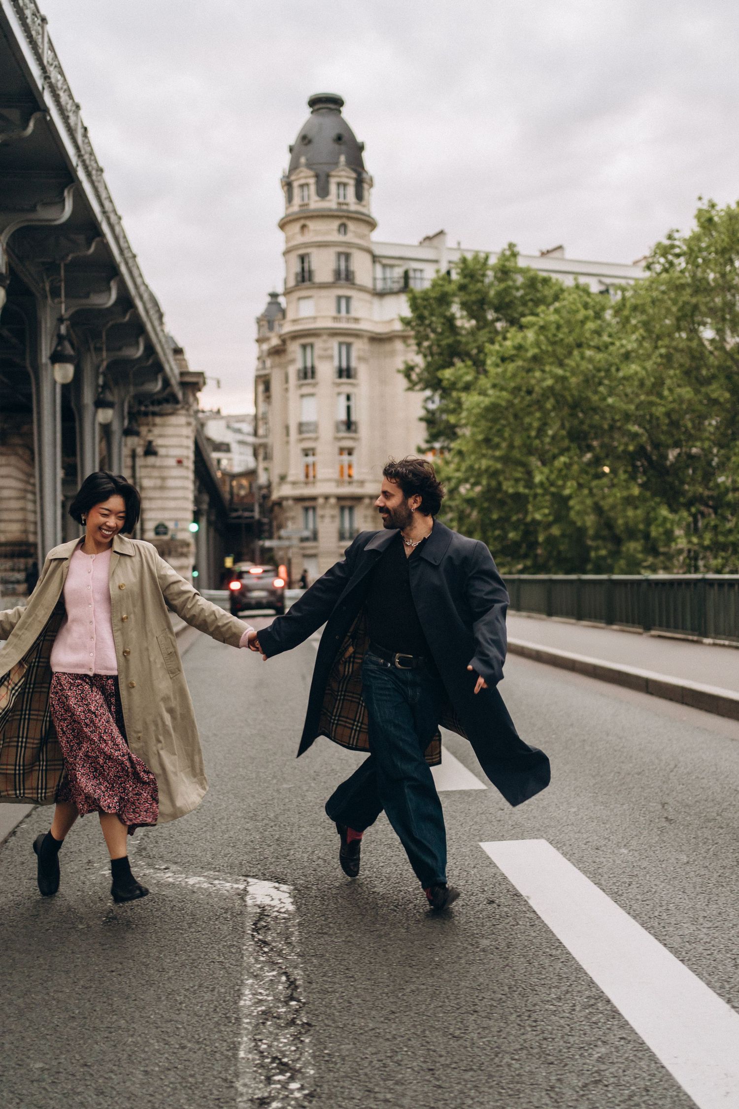 A joyful couple holds hands while crossing a Parisian street, dressed in stylish coats with classic Haussmann buildings behind them.