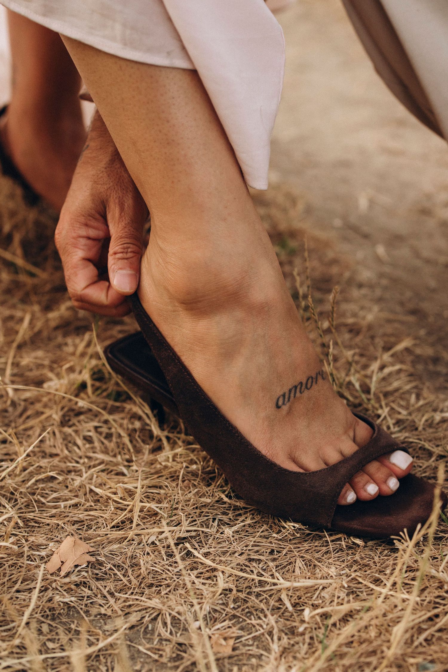 Close-up of a woman's foot with an amor tattoo, wearing a brown open-toe heel sandal on dry grass.