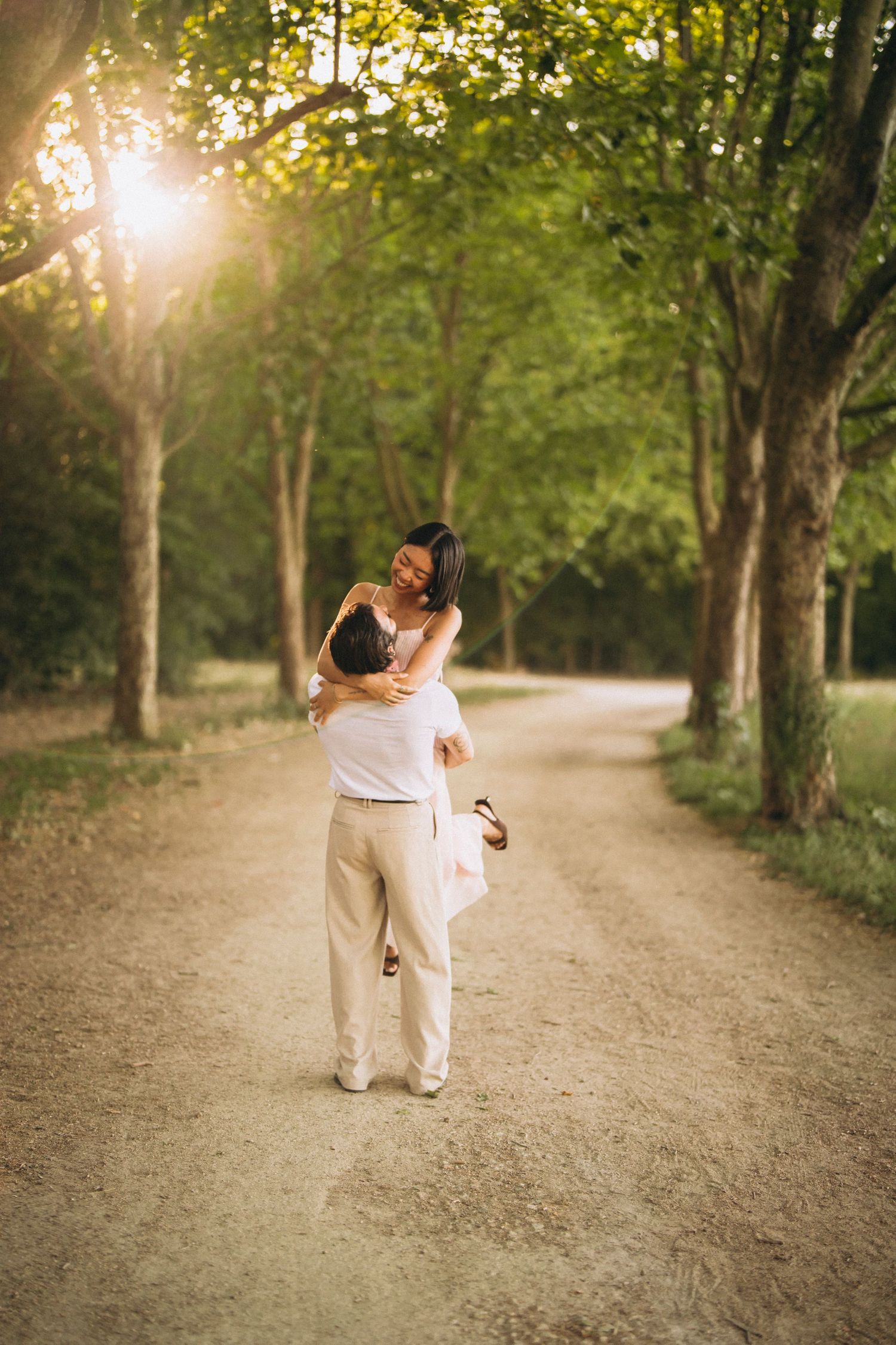 Couple embracing on a sunlit tree-lined path, man lifting woman in white dress, golden hour light filtering through green trees.
