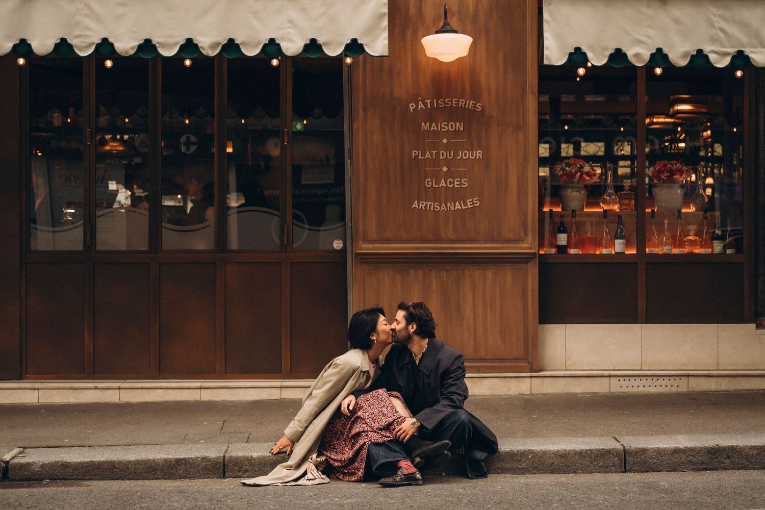 Romantic couple sharing a kiss while sitting on a cobblestone street outside a warmly lit Parisian patisserie at night.