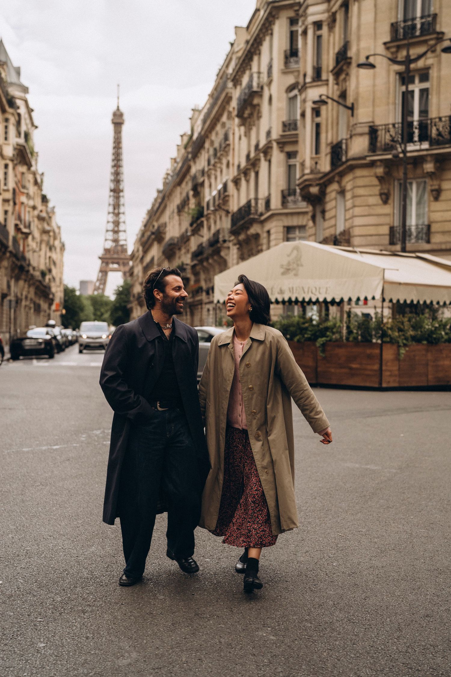 A couple walks and laughs together on a Parisian street with the Eiffel Tower visible in the background.